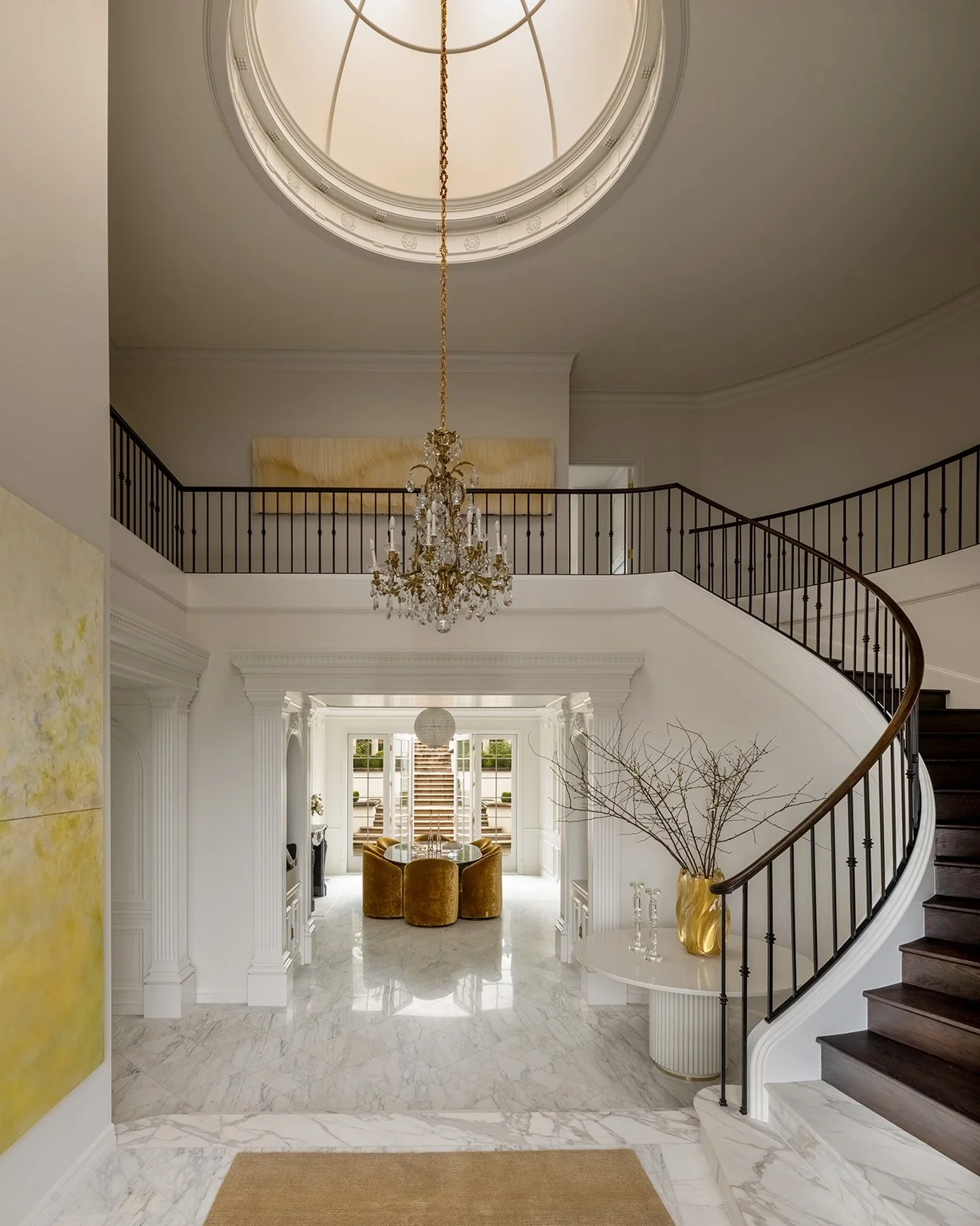 Interior view of a luxurious foyer with a curved staircase, chandelier, marble floors, and a sitting area with four yellow velvet chairs near large glass doors.
