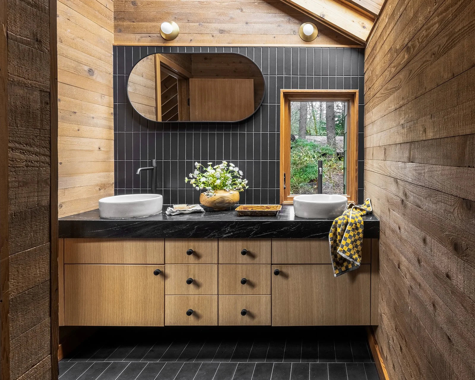 A rustic bathroom with wooden walls, a black tiled accent wall, a black countertop with two white vessel sinks, a round mirror, a window showing trees outside, a vase with flowers, and a yellow patterned towel. Rucon Build
