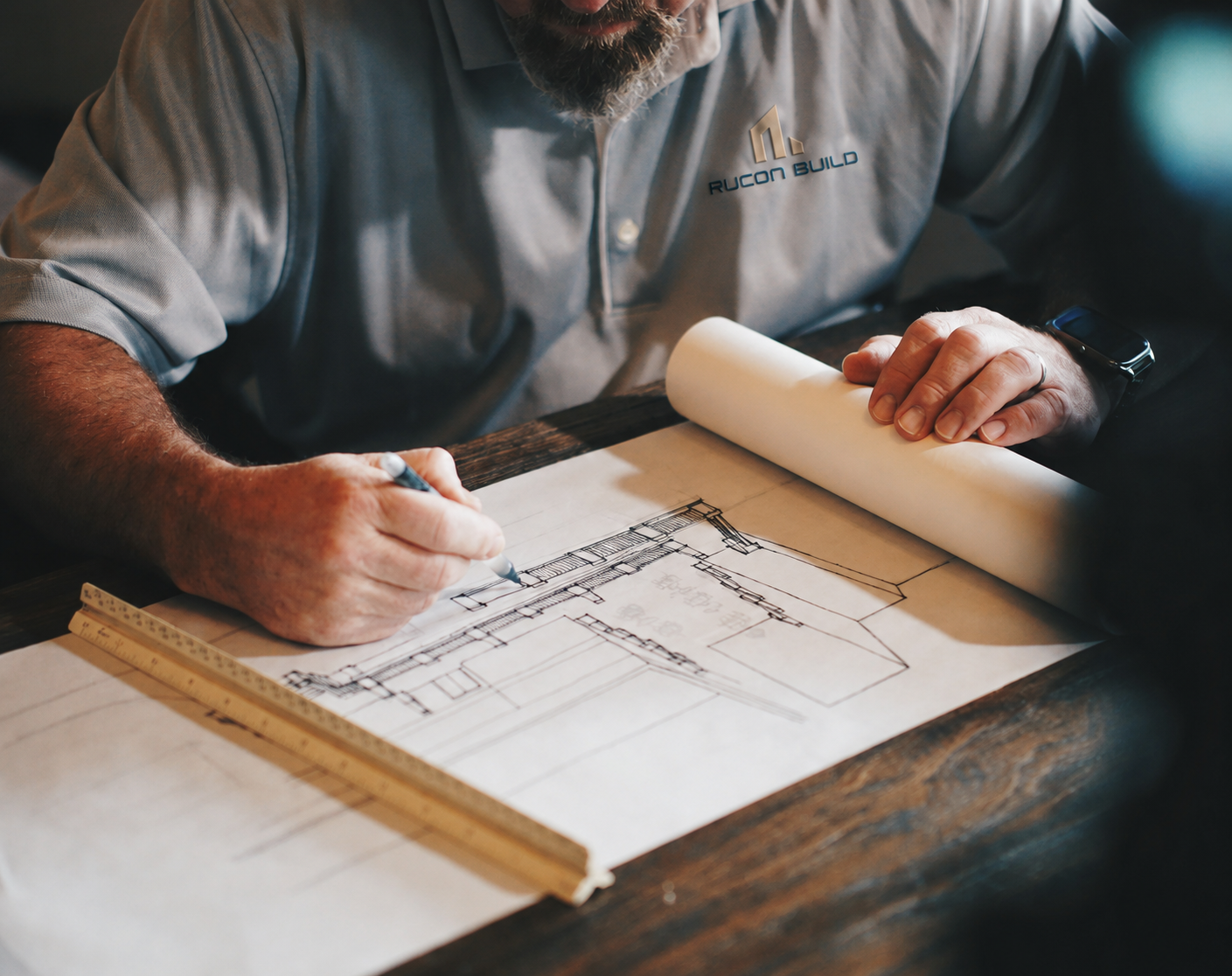A man wearing a gray shirt with a logo that says 'RUCON BUILD' is drawing architectural blueprints on large paper on a wooden table.
