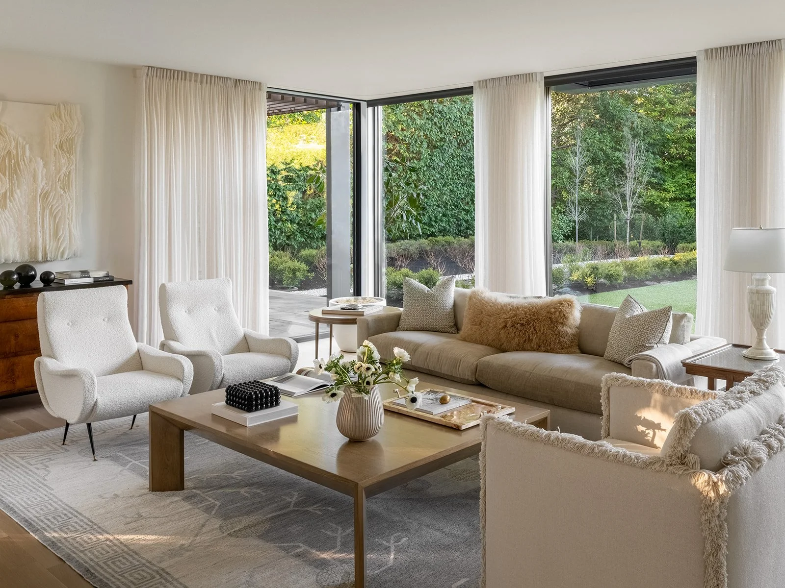 Bright living room with white curtains, beige sofa with throw pillows, white armchairs, wooden coffee table, and large windows showing greenery outside. Rucon Build Work