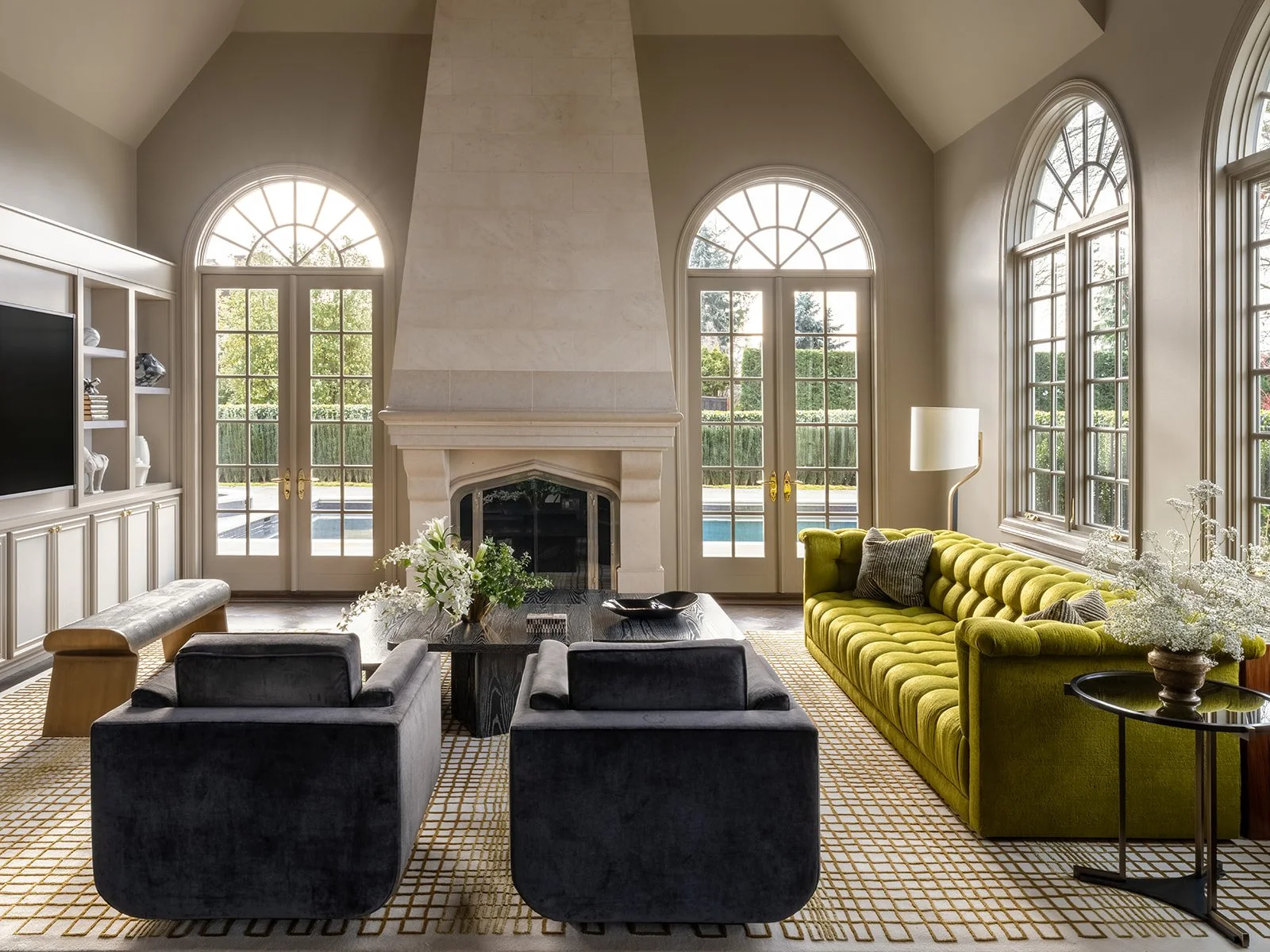 Living room with large arched windows, a fireplace, a green sofa, two dark velvet armchairs, and a black coffee table with floral arrangement.