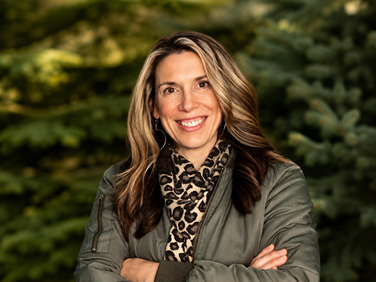 Smiling woman with wavy brown hair wearing a green jacket and leopard print scarf, standing outdoors with greenery in the background.