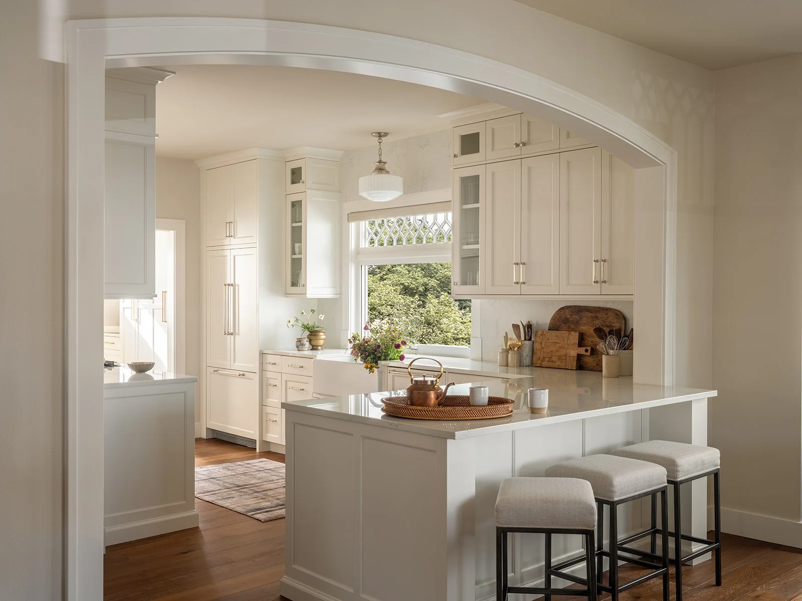 Bright white kitchen with cabinets, a central island, and a window view of greenery outside. Decorative items and kitchen utensils on the countertops, with three stools at the island.