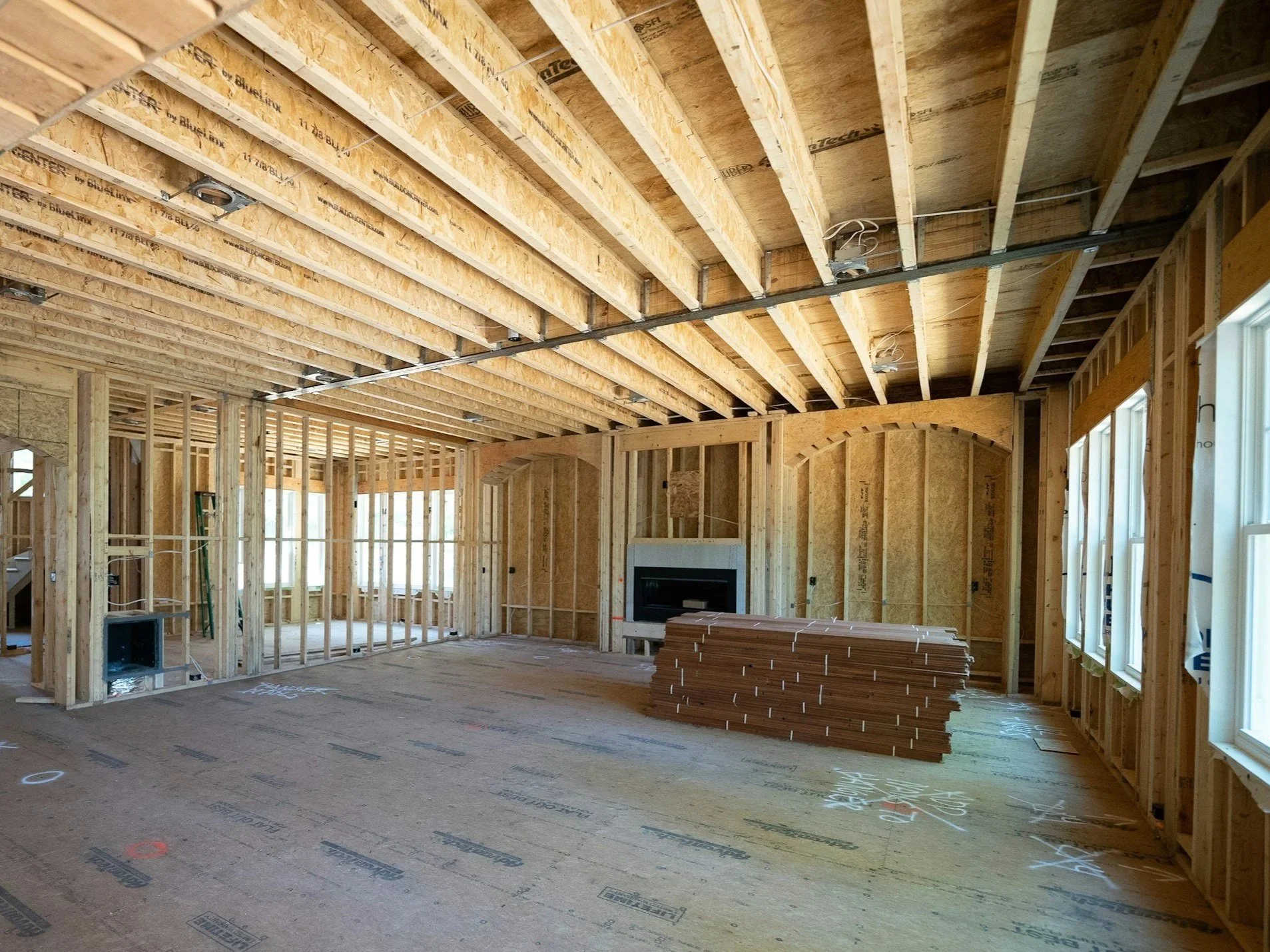 Interior of a house under construction with wooden framing, exposed ceiling beams, and windows along the side. Piles of wooden planks are stacked on the floor, and electrical wiring is visible.