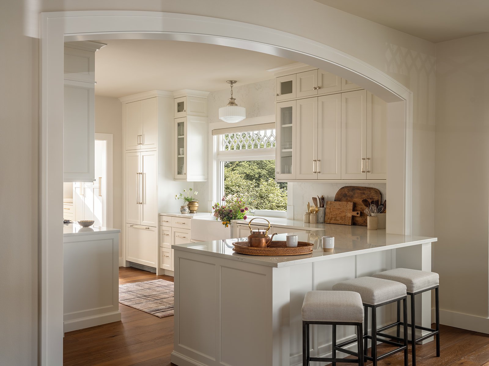 Bright kitchen with white cabinetry, a central island with a marble countertop, and a large window overlooking trees. Bar stools with white cushions are at the island. Decor includes a copper teapot, cups, potted plants, and wooden cutting boards.