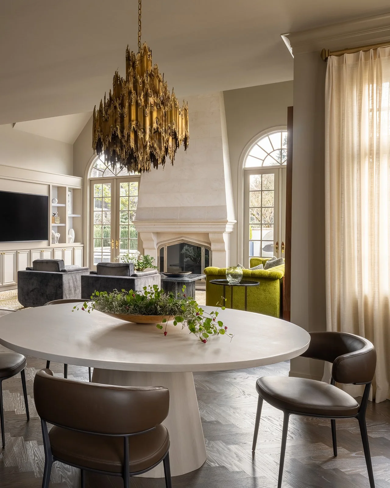 Living room with a coffee table, green and grey chairs, a fireplace, large windows, and a gold chandelier. Rucon Build
