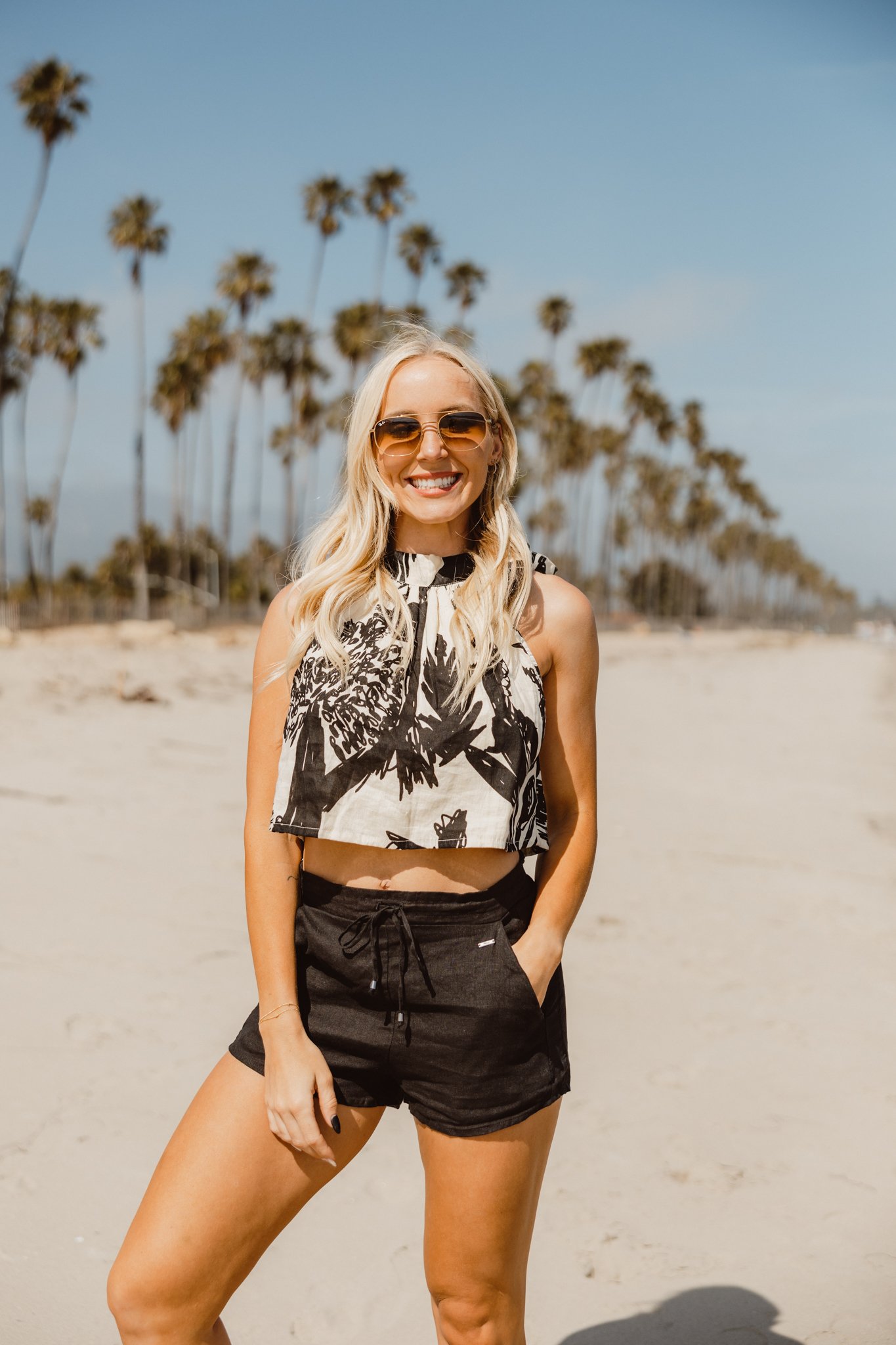 A smiling blonde woman wearing sunglasses, a black and white tropical print sleeveless crop top, and black shorts standing on a sandy beach with a row of tall palm trees in the background under a clear blue sky.