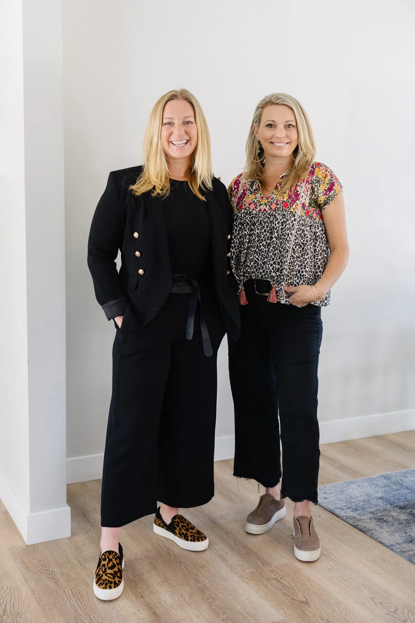 Two women standing next to each other indoors against a plain white wall, smiling at the camera. The woman on the left is wearing a black blazer and matching pants, with leopard print slip-on shoes. The woman on the right is wearing a colorful, patterned top with black pants and beige slip-on shoes.