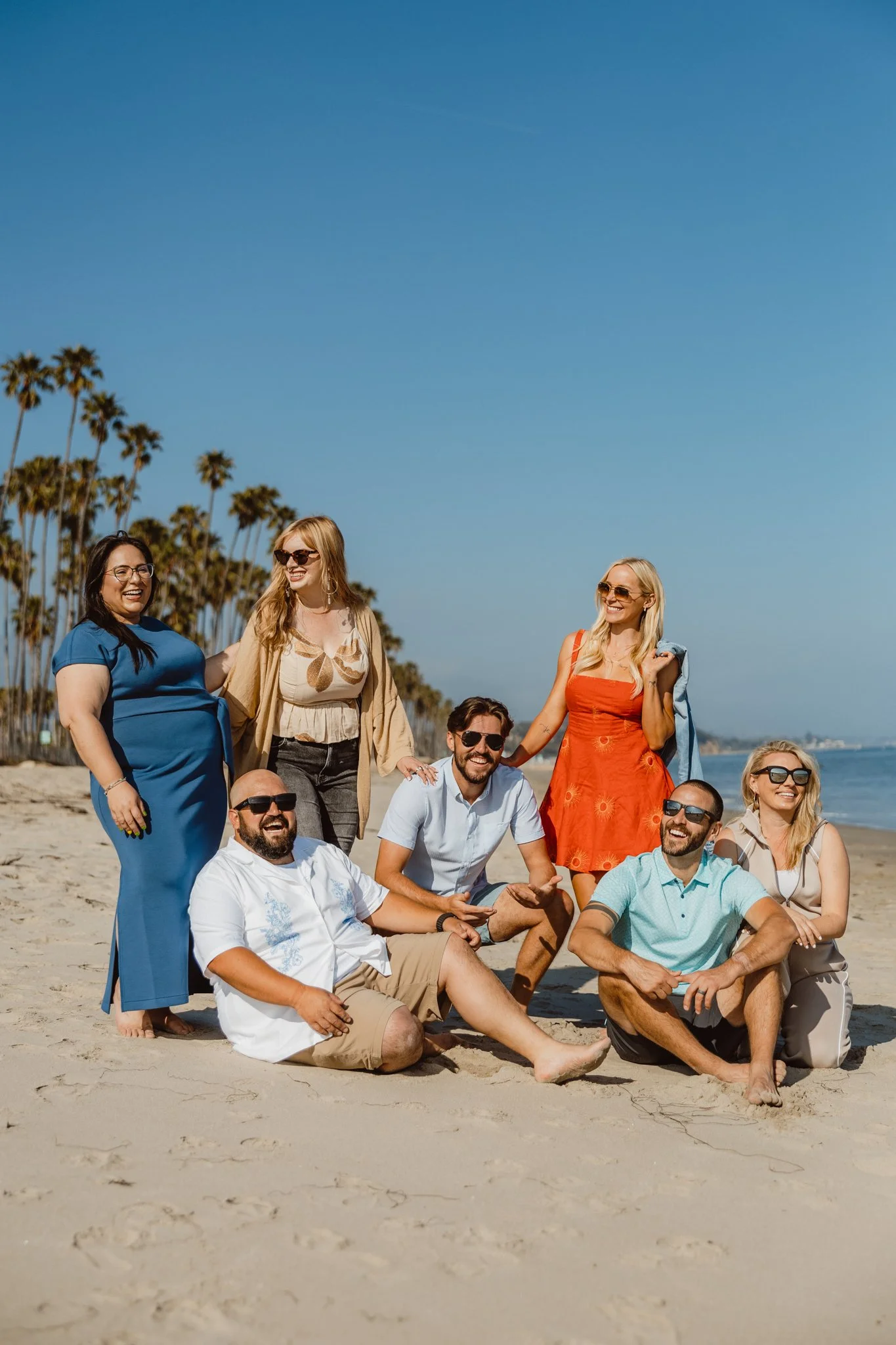 A group of smiling friends posing on a sandy beach with palm trees and blue sky in the background. Some are sitting, some are standing, and they are dressed in casual summer clothing.