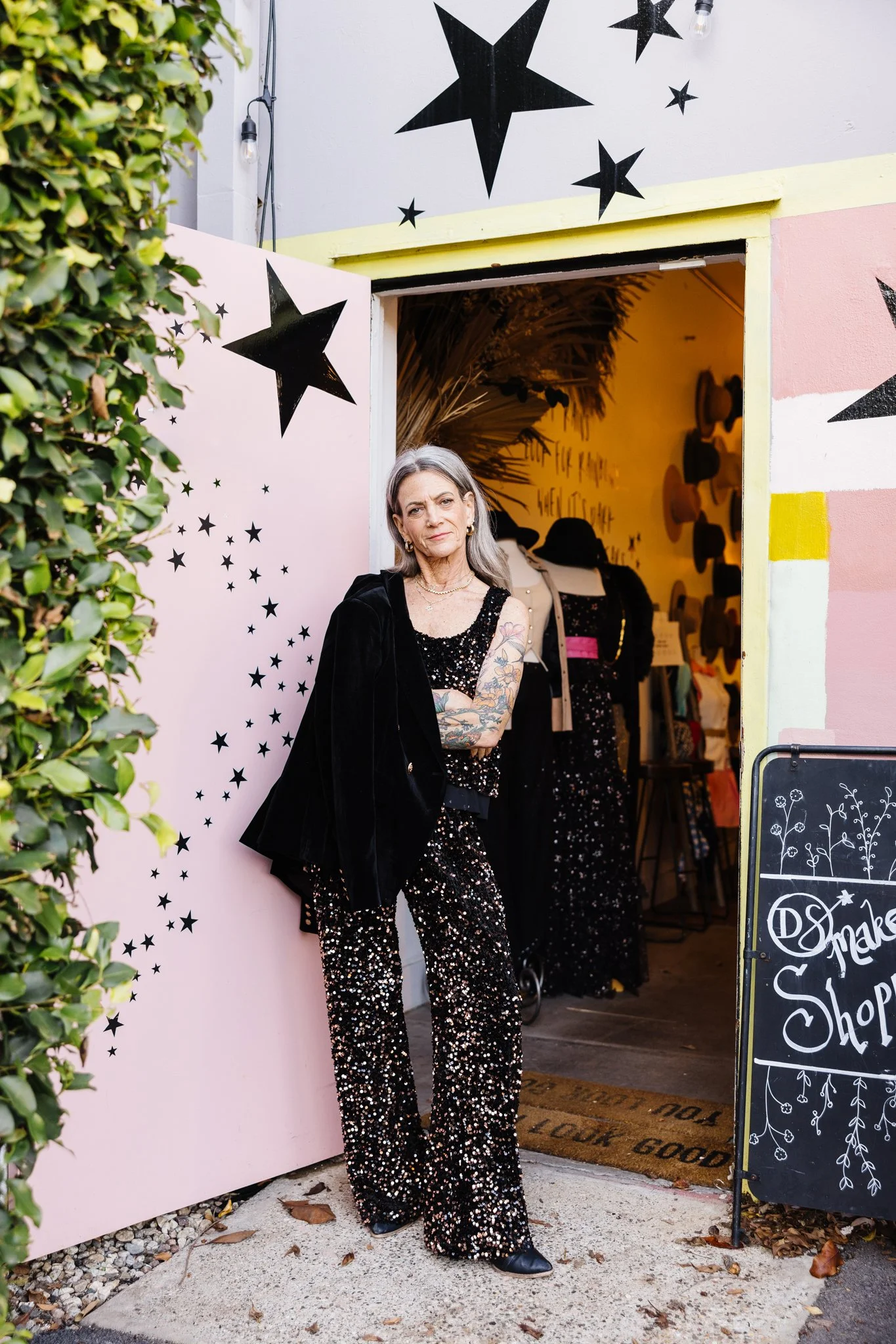 A woman with gray hair and tattoos on her arms stands in front of an entrance to a store with black star decorations on the wall. She is wearing a black glittery outfit and black shoes, with her arms crossed and a serious expression.