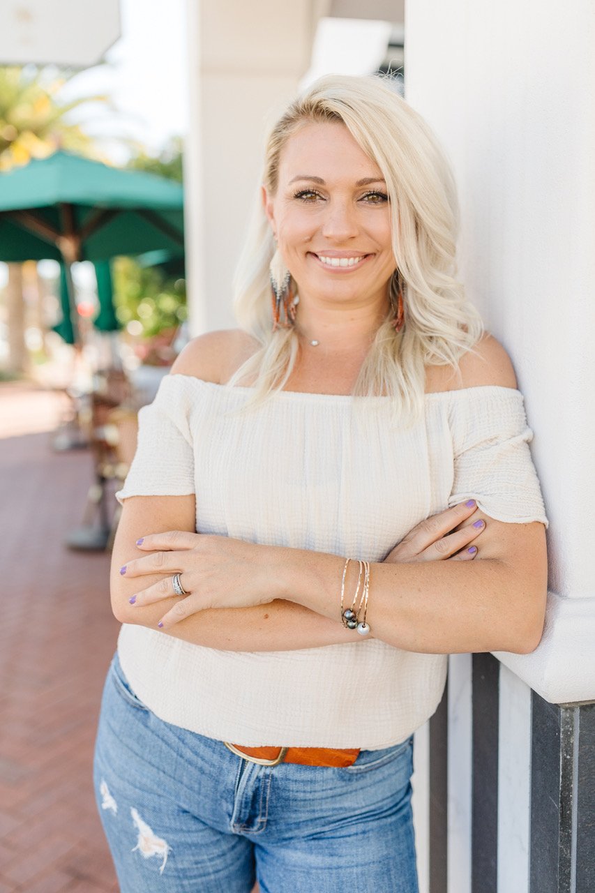 A blonde woman with wavy hair leaning against a wall outdoors, smiling at the camera. She's wearing a white off-shoulder top, distressed blue jeans, and earrings.