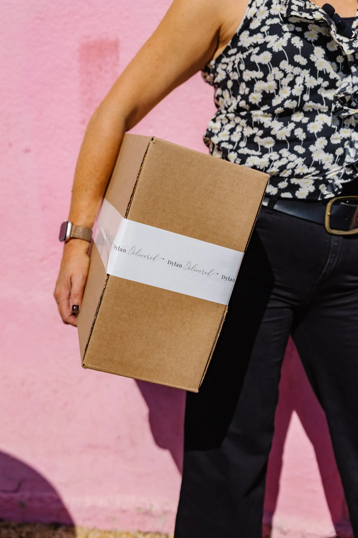 Person wearing a daisy print shirt holding a cardboard box labeled 'Dylan Delivered' against a pink background.