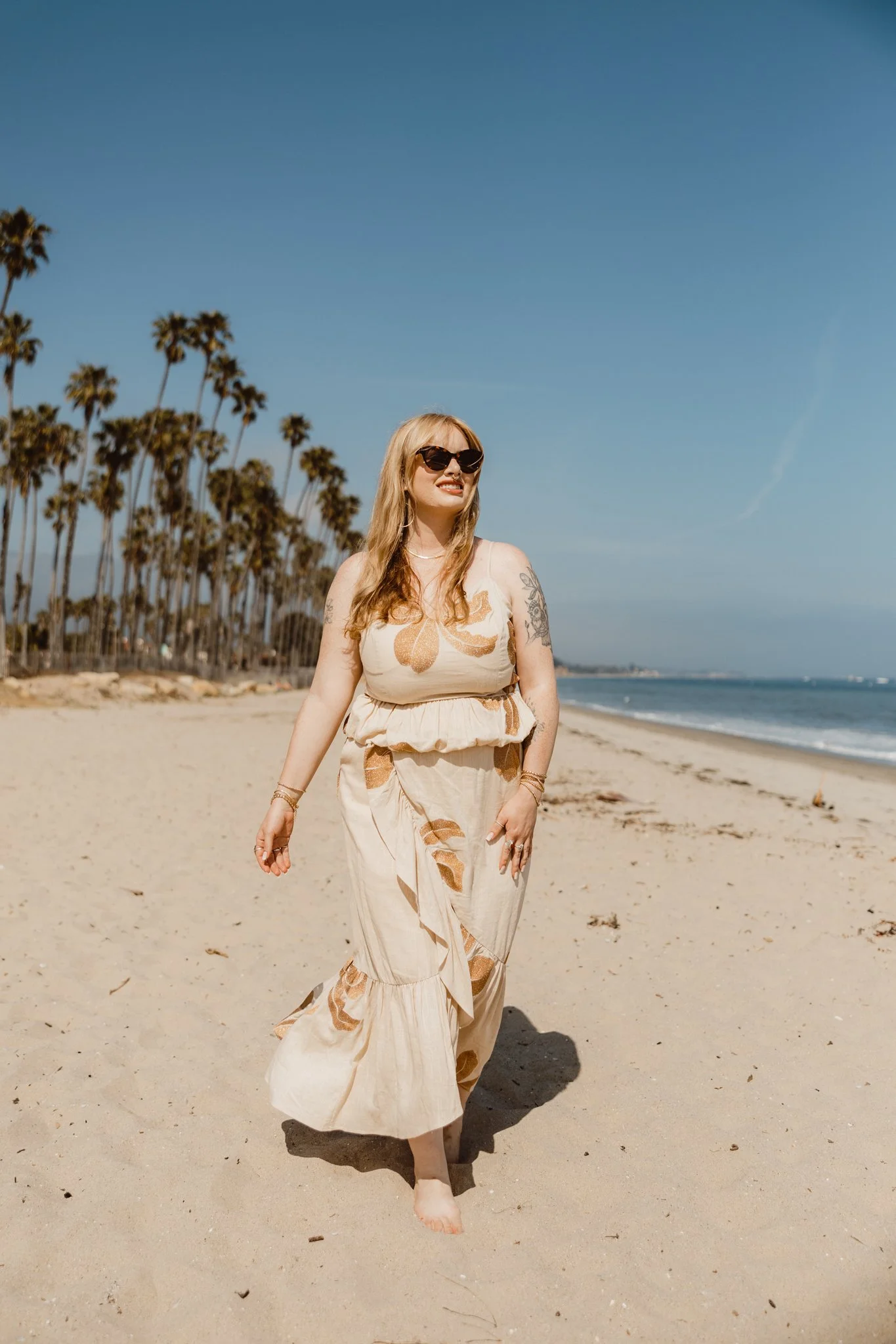A woman wearing sunglasses and a flowy dress walking barefoot on a sandy beach with palm trees and the ocean in the background.