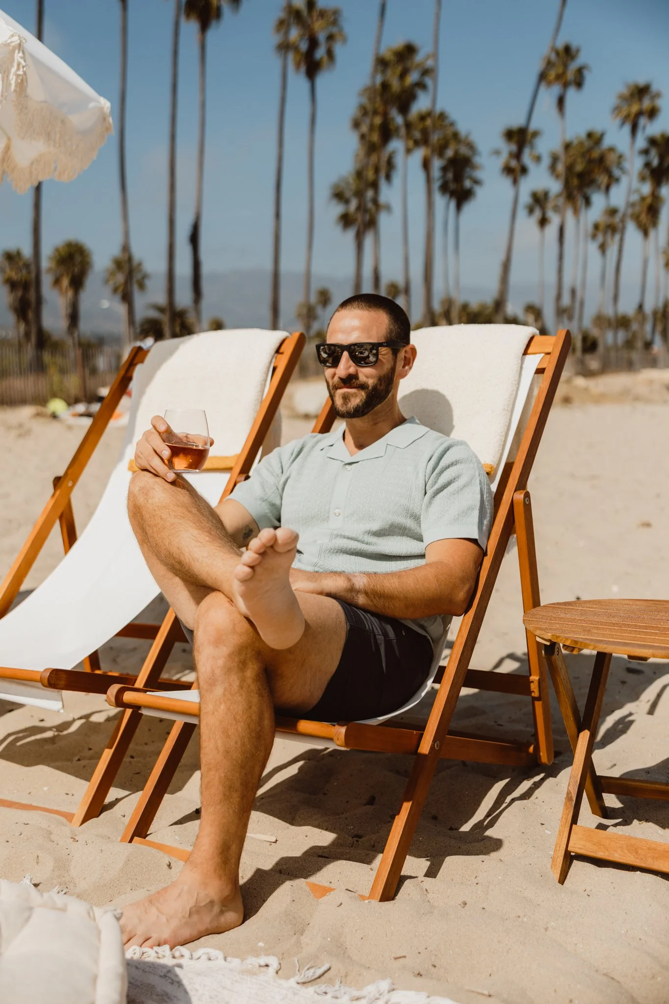 A man relaxing on a lounge chair at the beach, wearing sunglasses and holding a glass of rosé wine, with palm trees and mountains in the background on a sunny day.