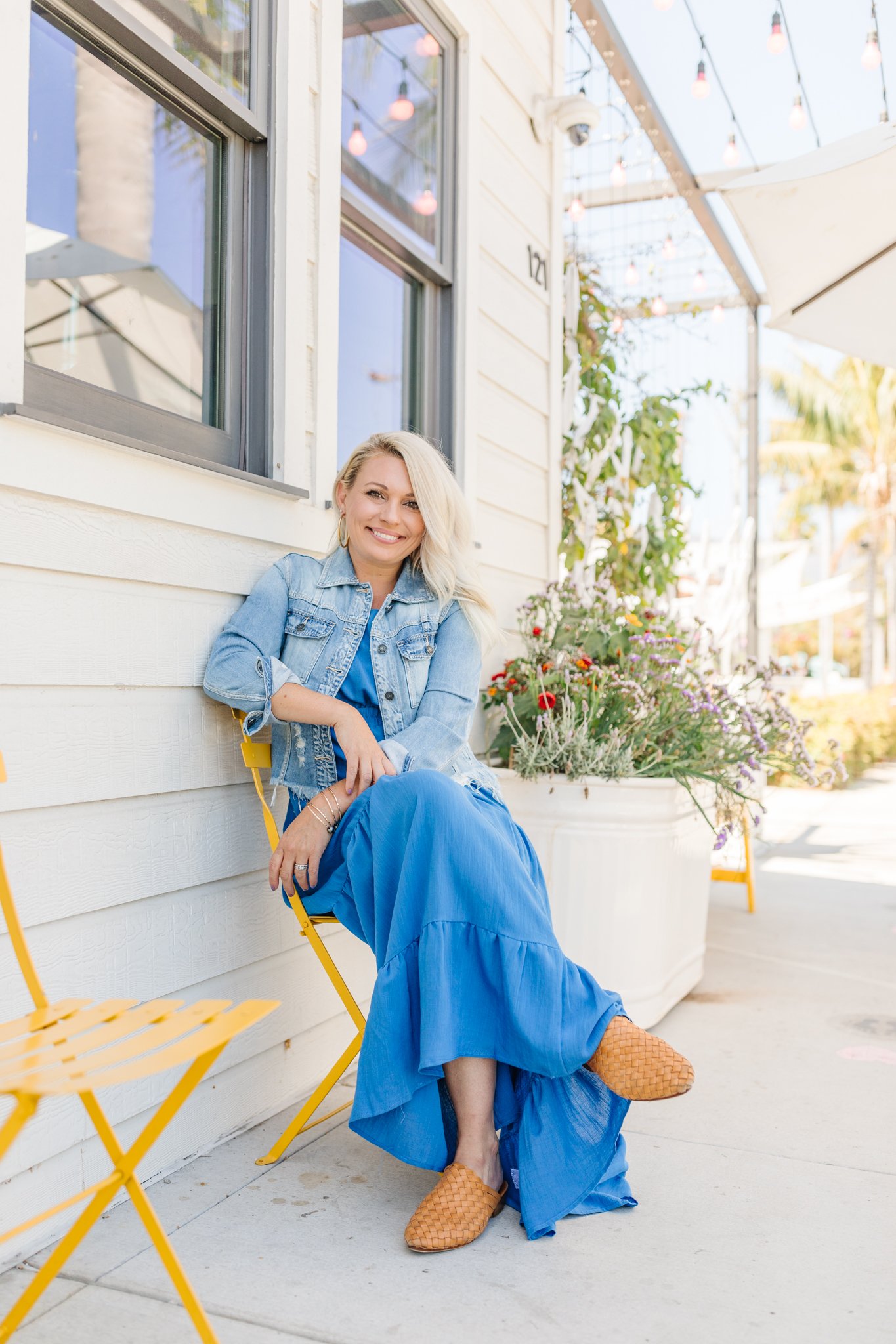 A woman with blonde hair wearing a denim jacket and a bright blue dress, smiling while sitting on a yellow chair outside a white house with large windows. She is also wearing tan woven slip-on shoes. There are potted flowers and string lights above.
