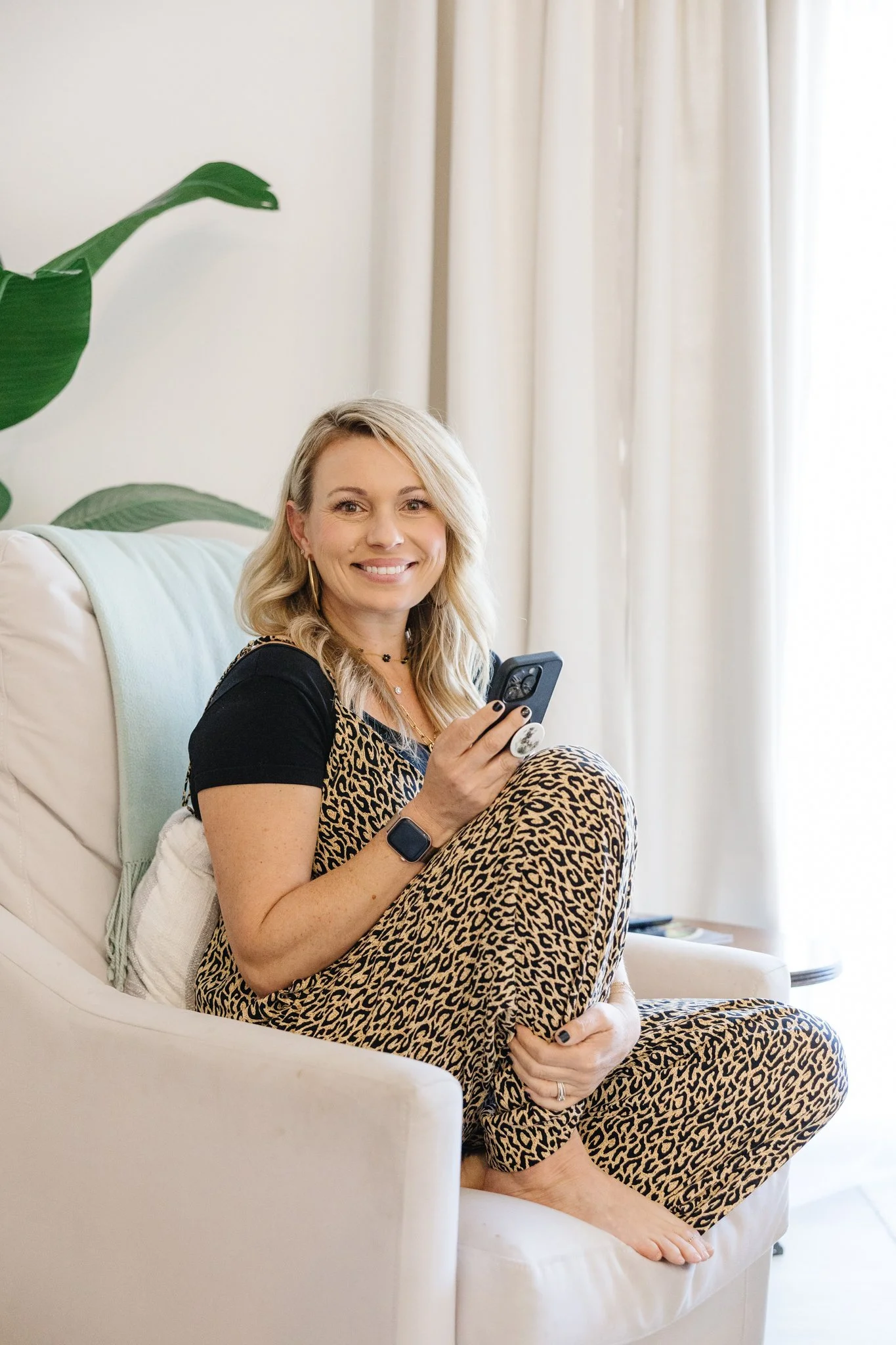 A woman with blonde hair sitting on a white armchair, holding a smartphone, wearing leopard print pants and a black top, smiling at the camera.