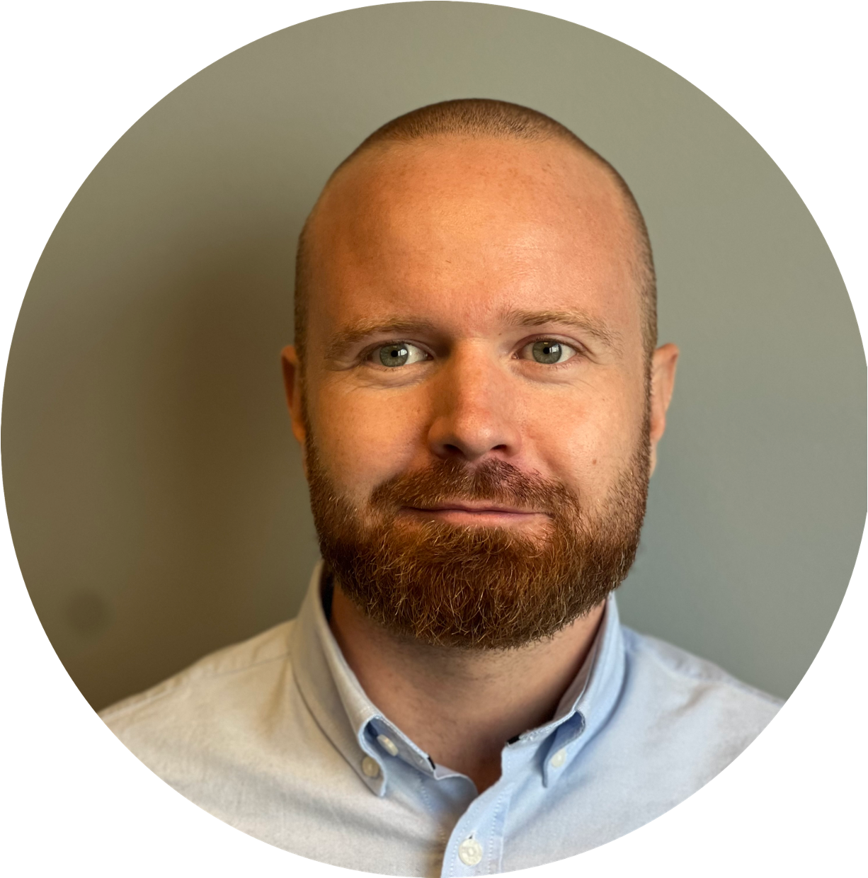 Portrett av en mann med kort hår og skjegg, iført lys blå skjorte, stående foran en grå vegg.