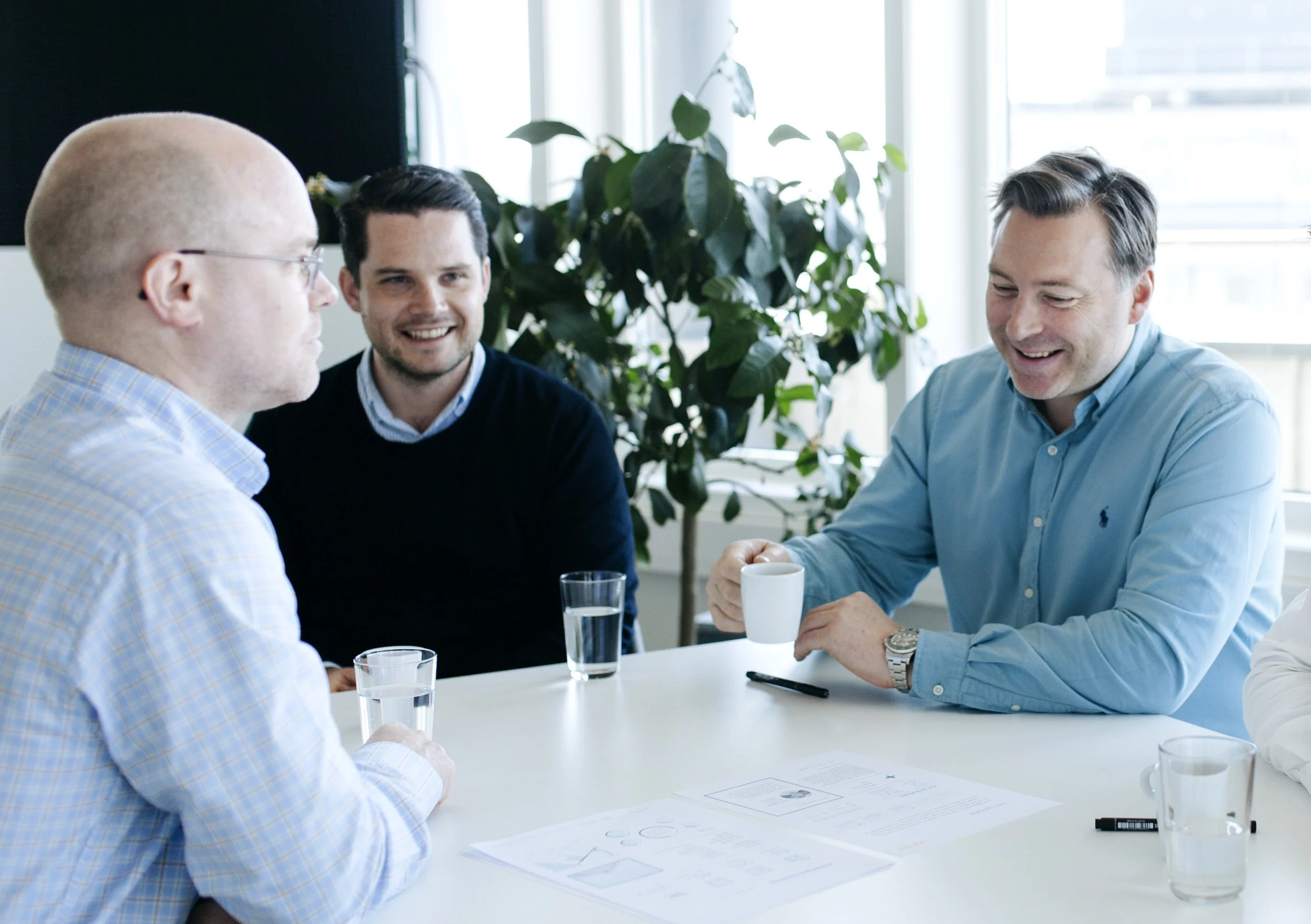 Tre menn i samtale rundt et bord med dokumenter, kopper og glass med vann, i et møtelokale med store vinduer og grønne planter.