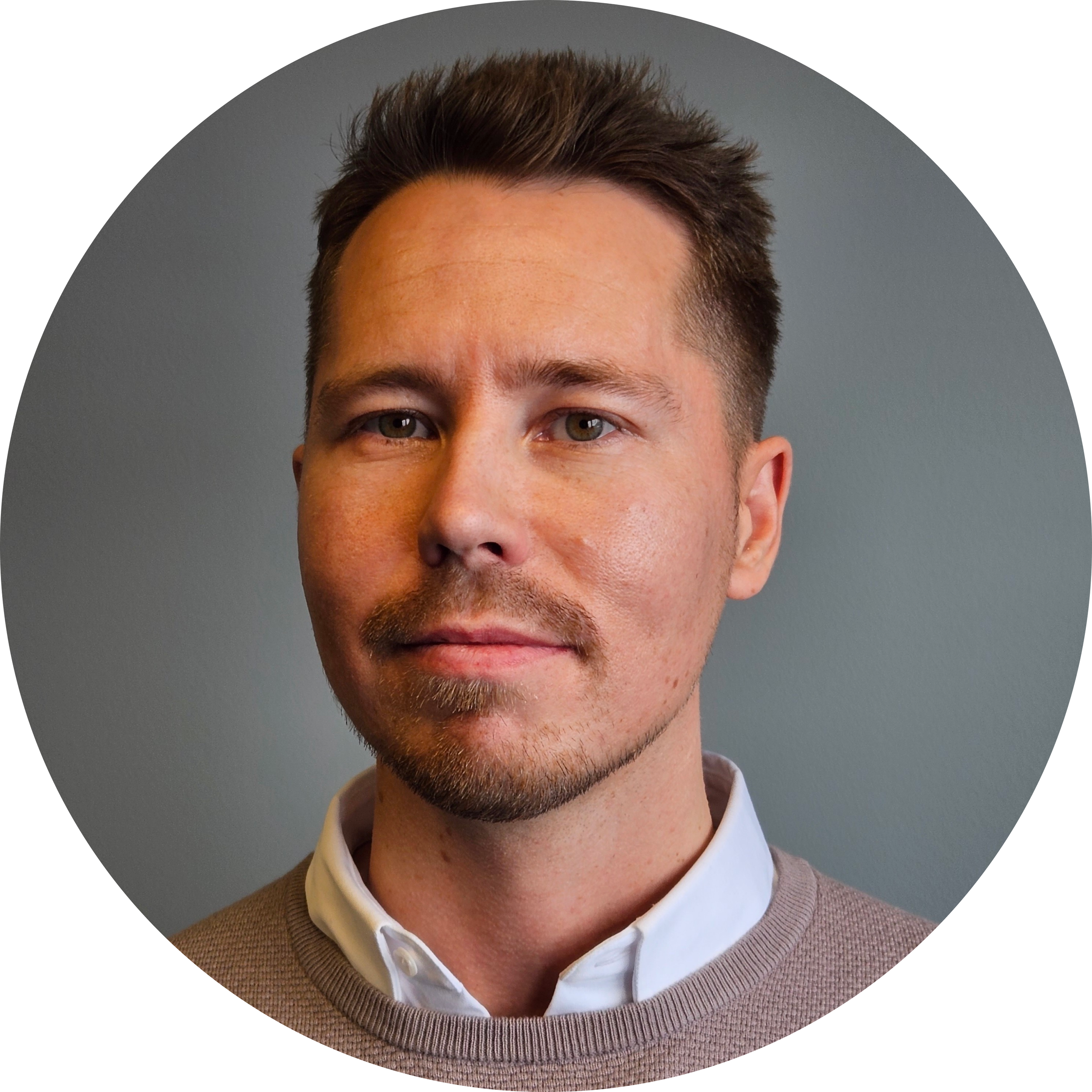 Porträtt av en man med mörkt, stylet hår, ljus hud och gröna ögon, klädd i en beige tröja och vit skjorta.