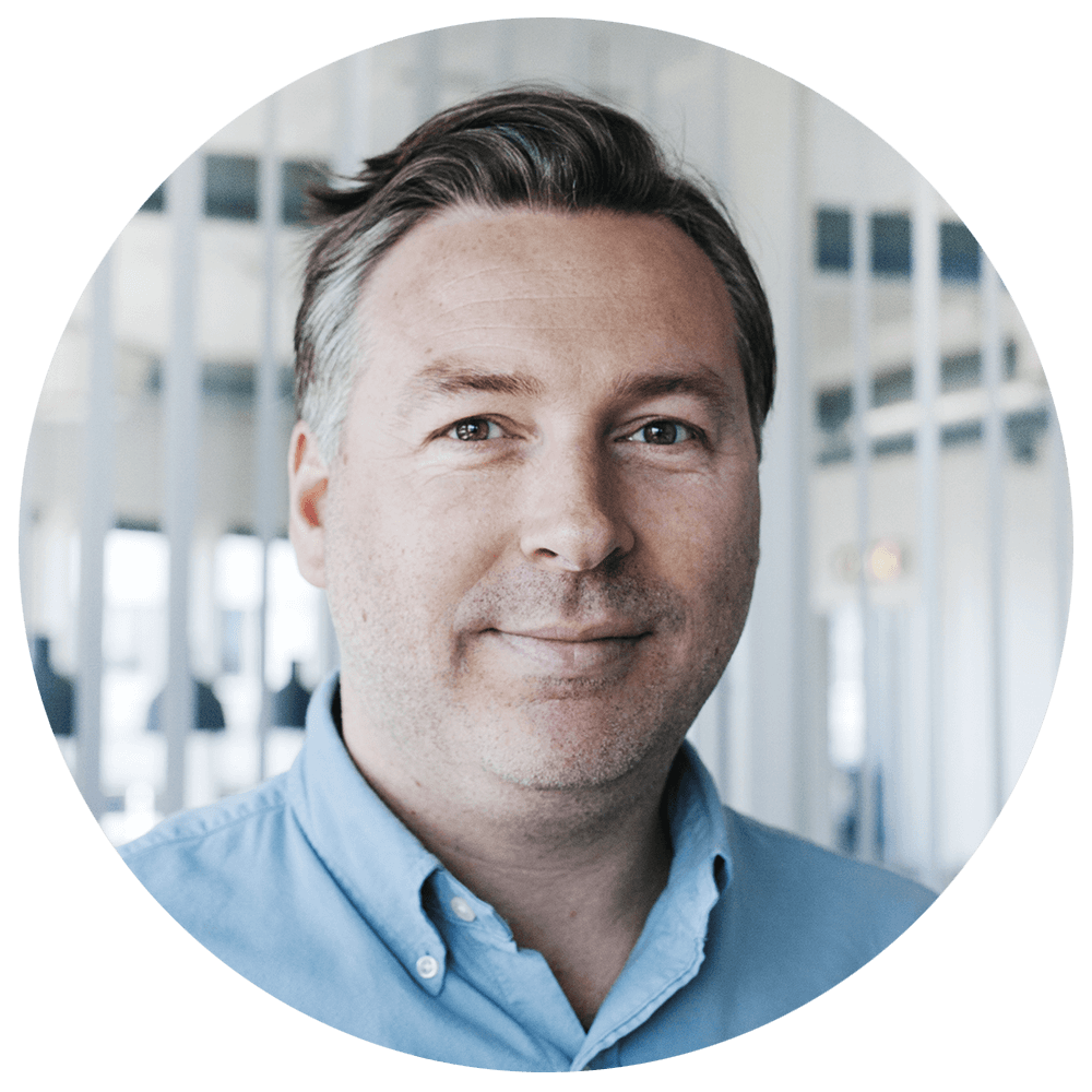 Portrett av en smilende mann med blå skjorte i innendørs miljø.