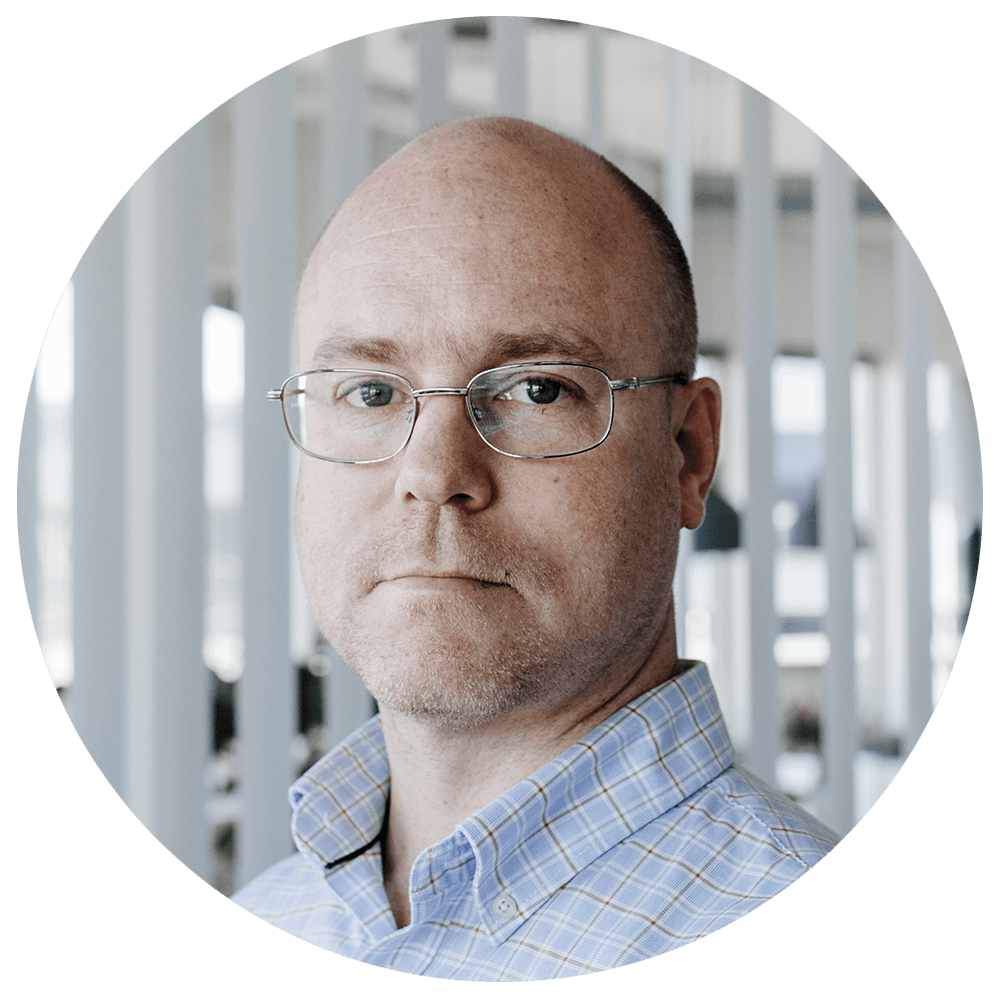 Portrett av en mann med briller og lyst skjorte foran en moderne bakgrunn med hvite vertikale stolper.