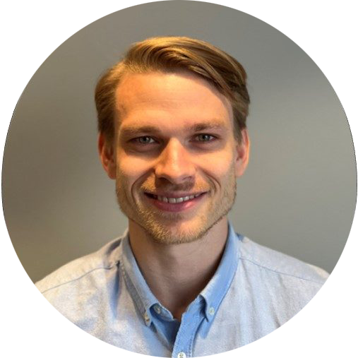 Et portrett av en mann med lyst hår, skjegg, iført en lys blå skjorte, smilende.