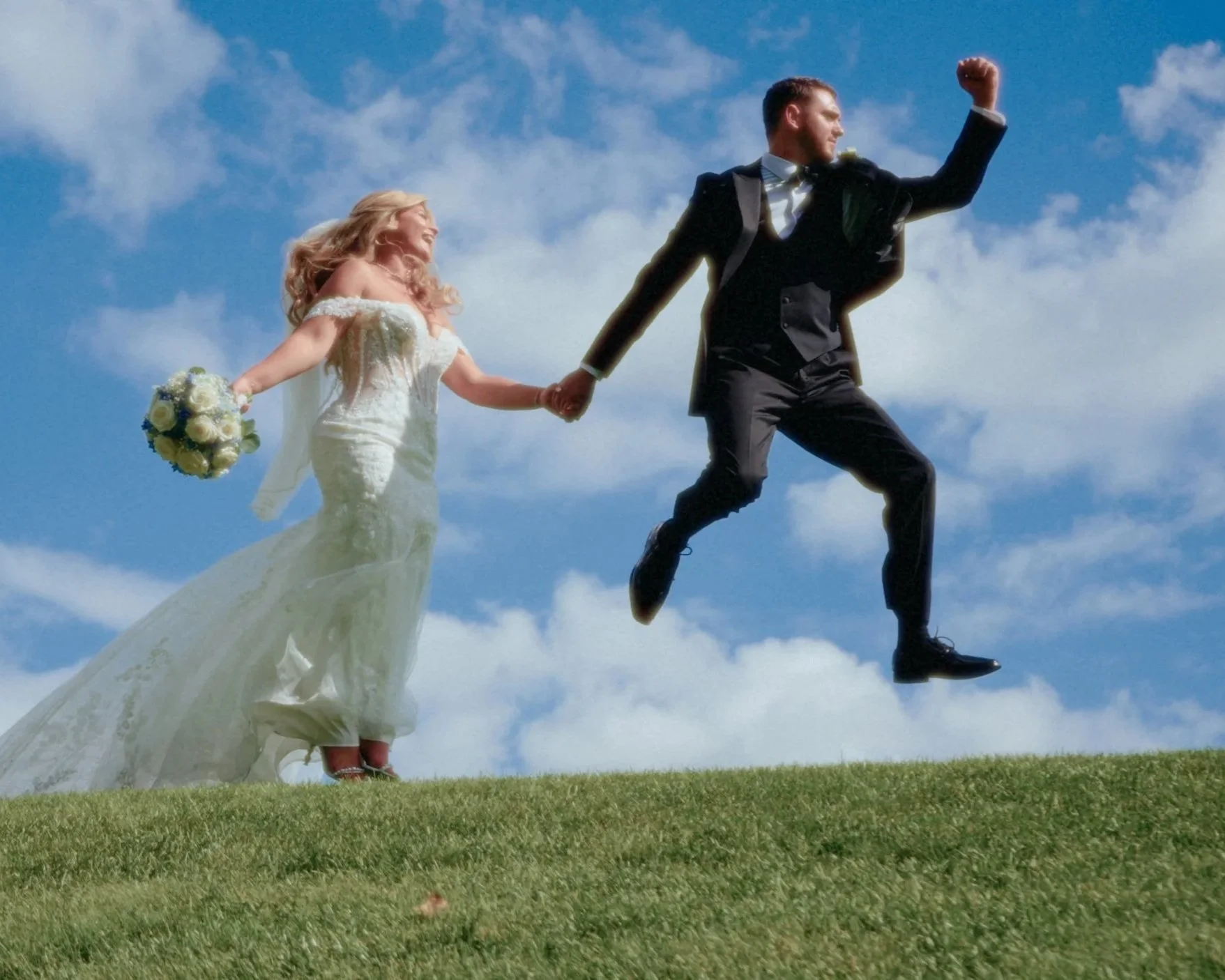 A bride and groom holding hands, with the groom jumping in the air while the bride watches and smiles, outdoors against a blue sky with clouds.
