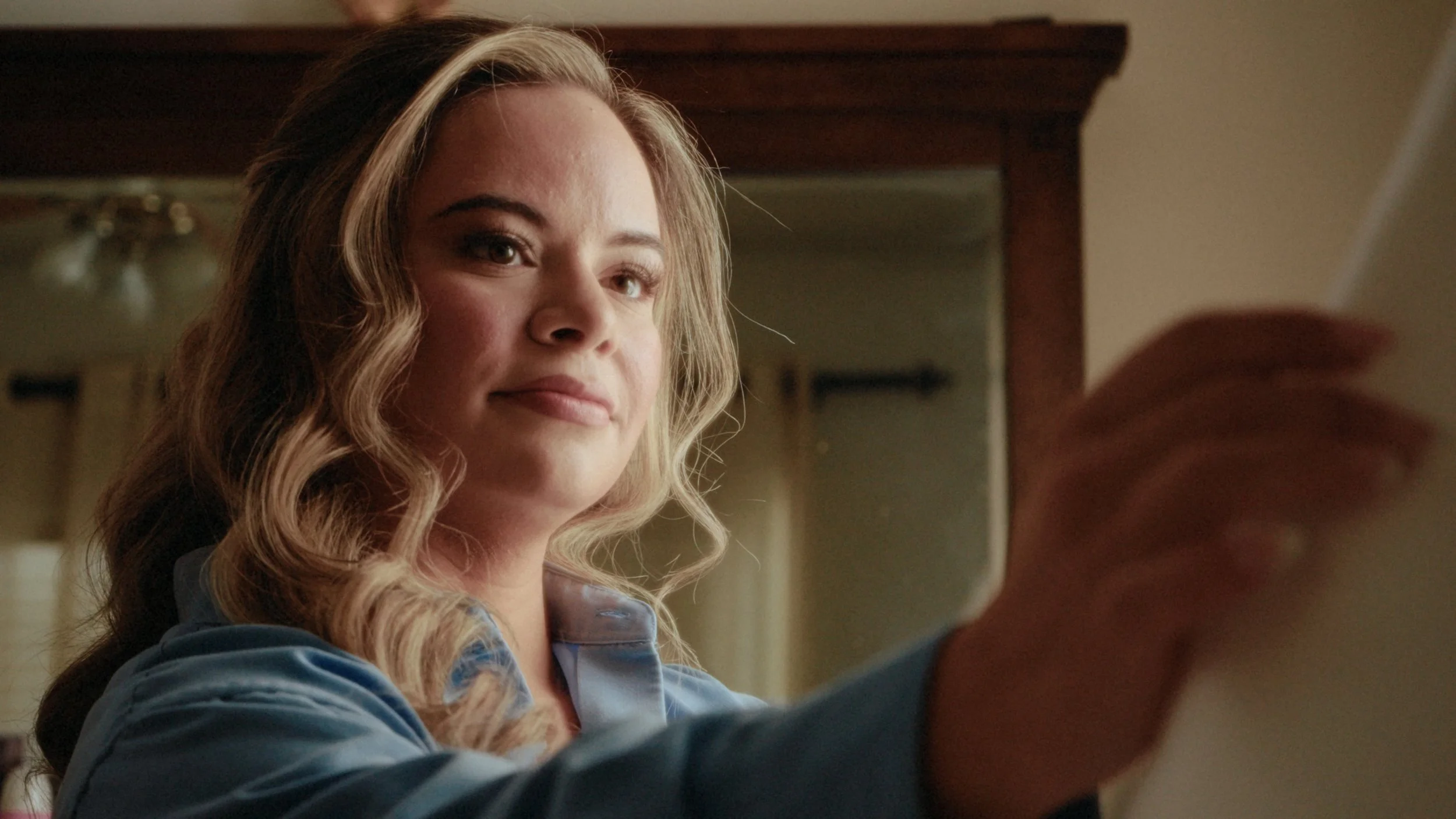 A woman with wavy blonde hair looking at a smartphone with a subtle smile.