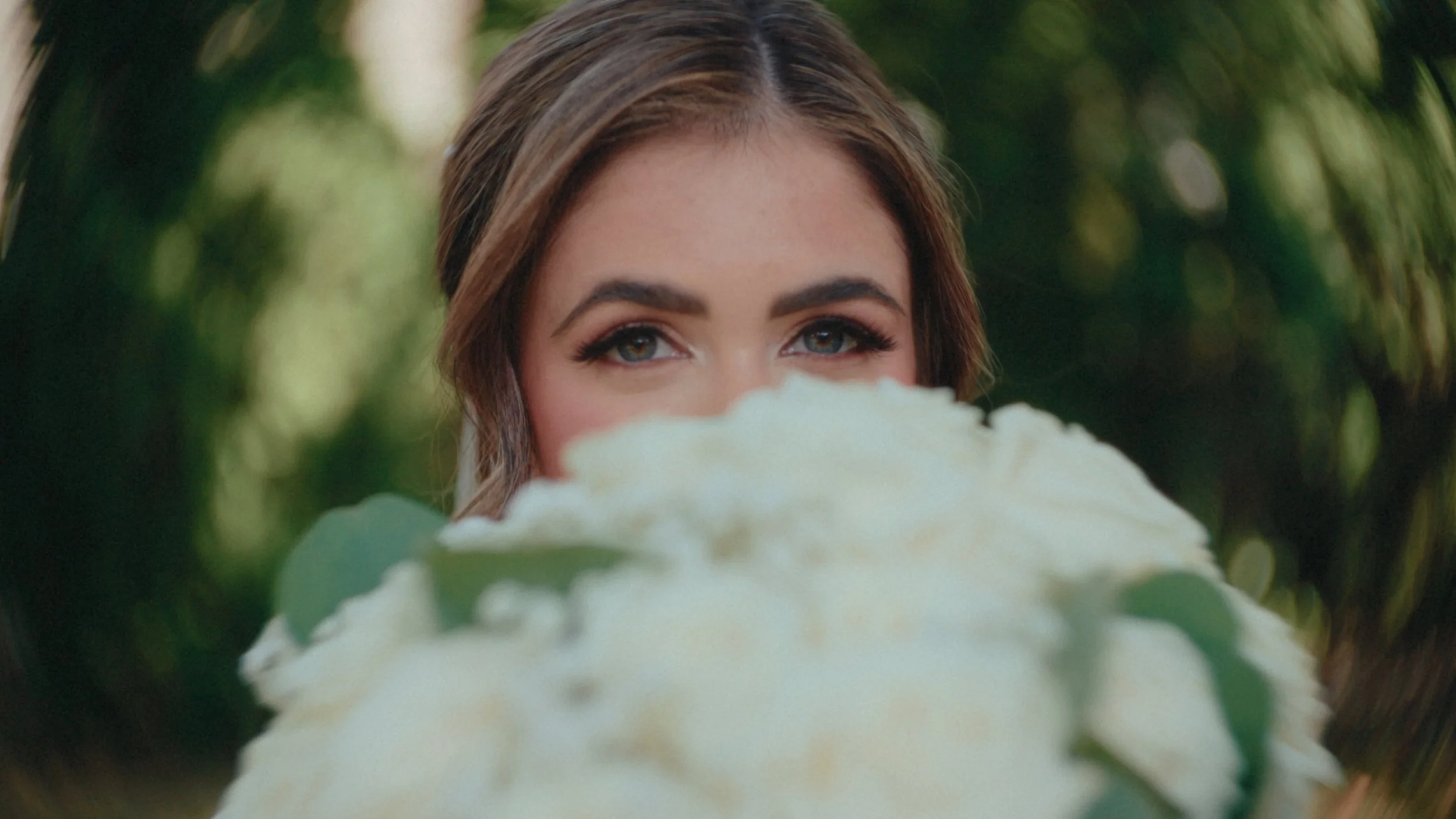Close-up of a woman with brown hair and blue eyes holding a large white flower close to her face, with green foliage in the background.