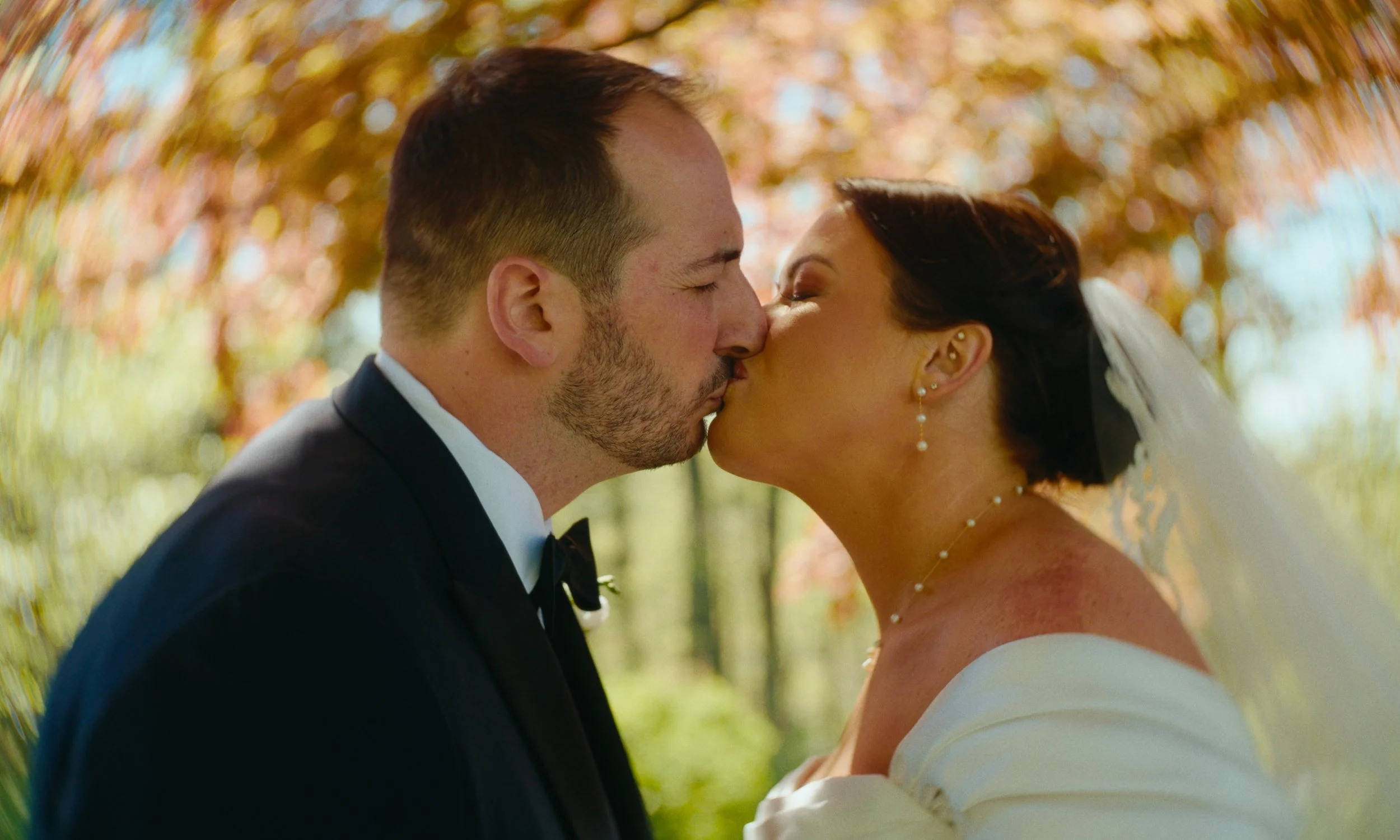 A couple dressed in wedding attire sharing a kiss outdoors with colorful autumn leaves in the background.