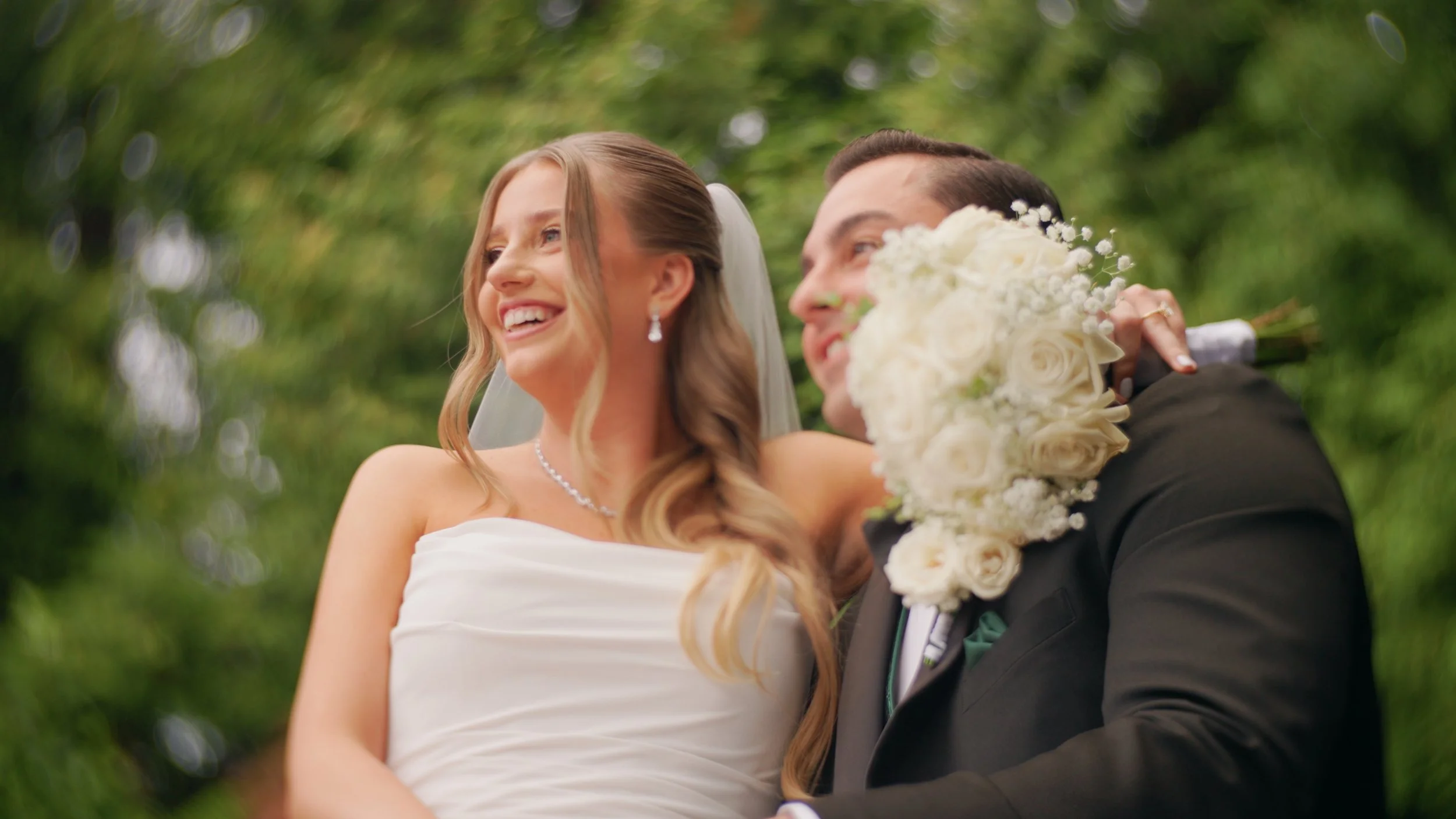 A happy bride and groom on their wedding day, smiling and looking joyful outdoors with a green, blurred background.