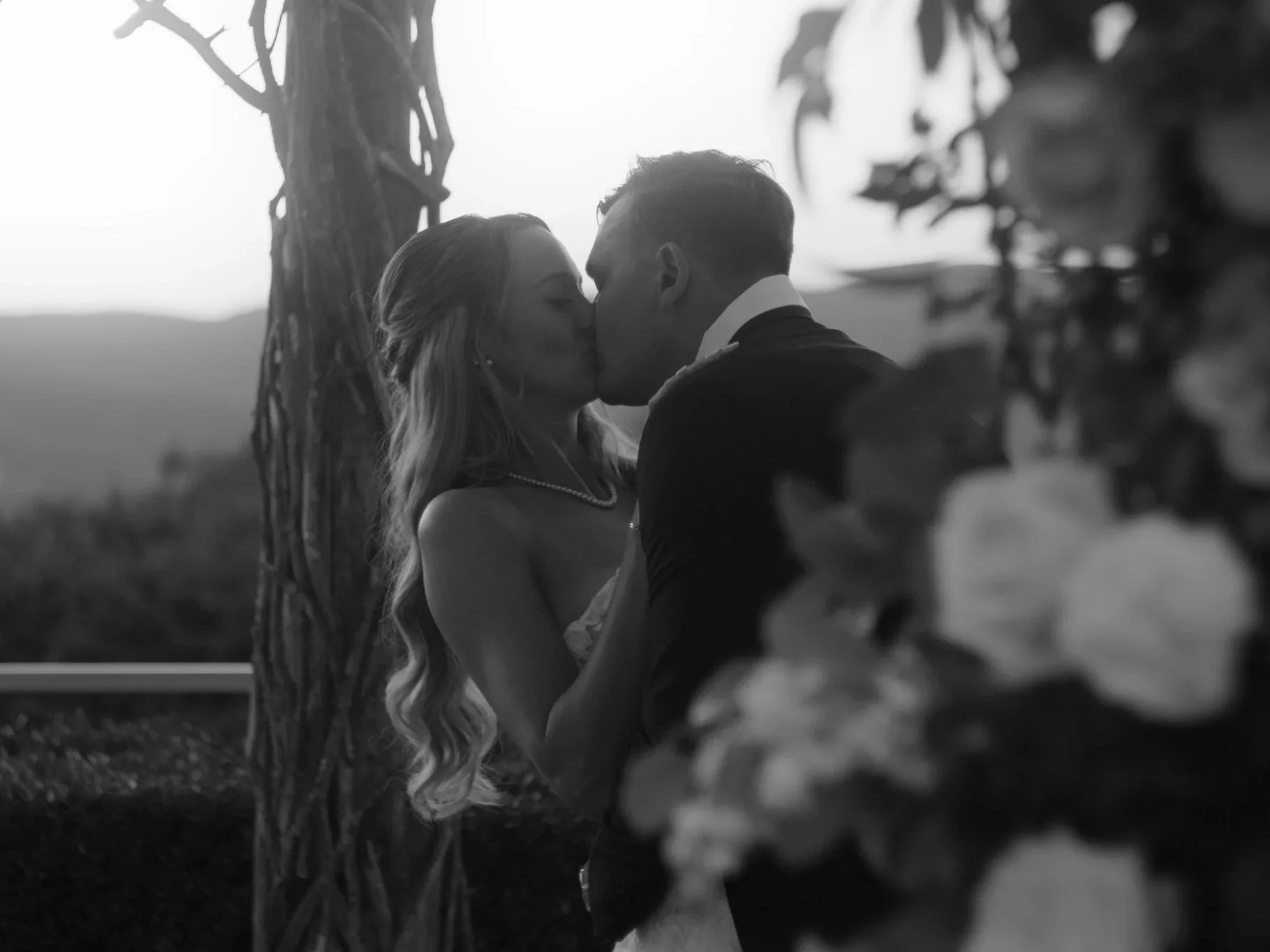 A black and white photo of a man and woman kissing outdoors near a tree, with blurred foliage in the foreground.