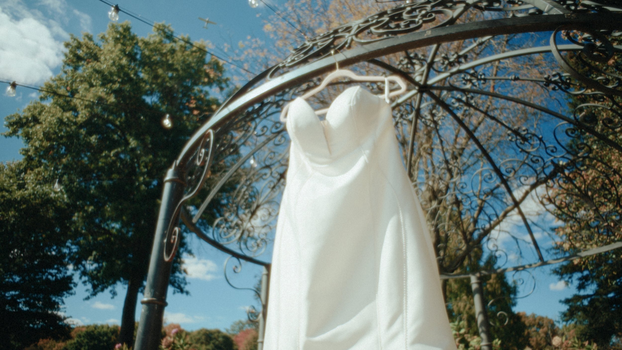 White dress hanging on a decorative metal arch outdoors with trees and blue sky in the background.