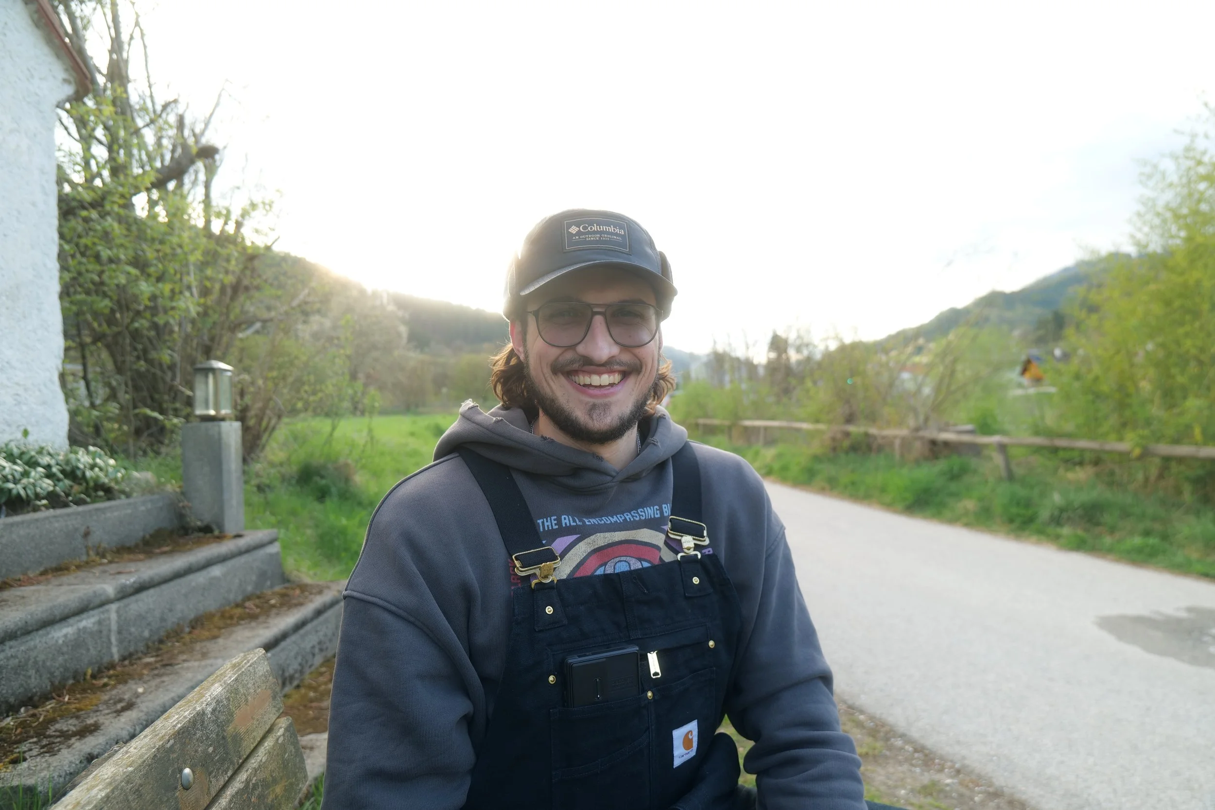 Lächelnder junger Mann mit Brille, dunklem Hoodie und schwarzer Carhartt-Weste, sitzt auf einer Parkbank im Freien, mit Bäumen und Hügel im Hintergrund, Sonnenlicht hinter ihm.