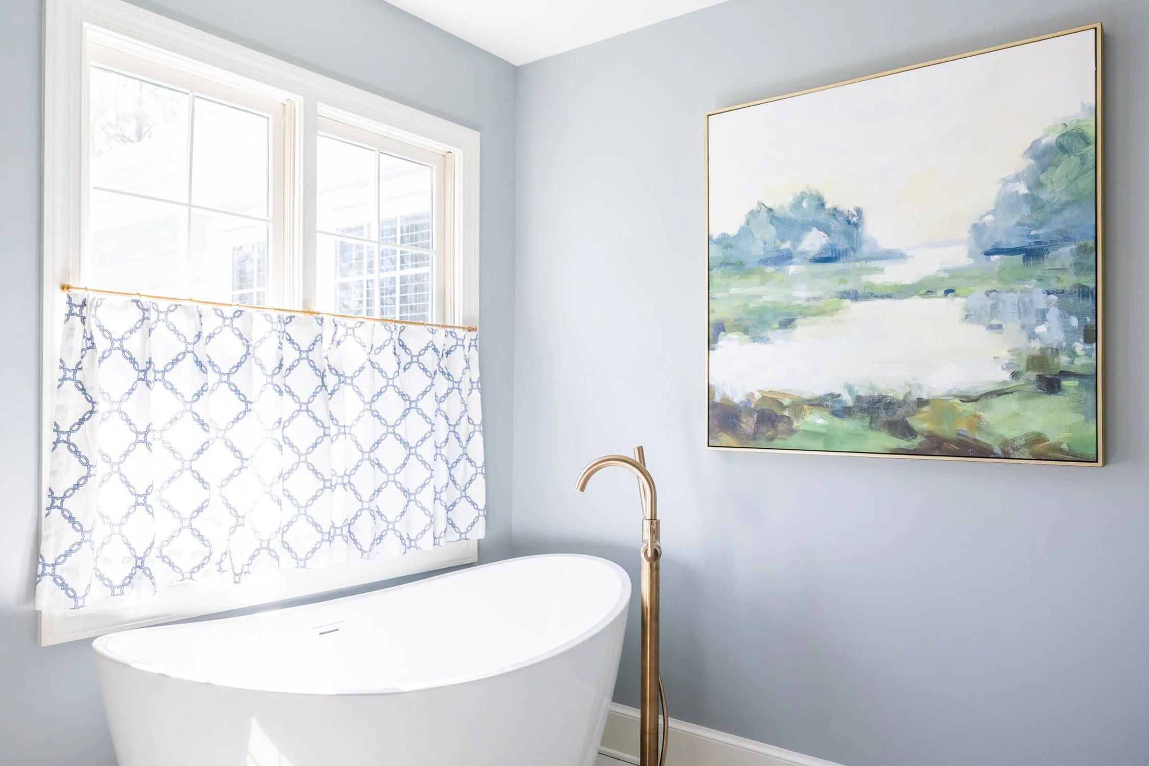 A bathroom with large windows, a white bathtub, a gold freestanding faucet, a framed landscape painting on the wall, and a white shower curtain with a blue pattern.