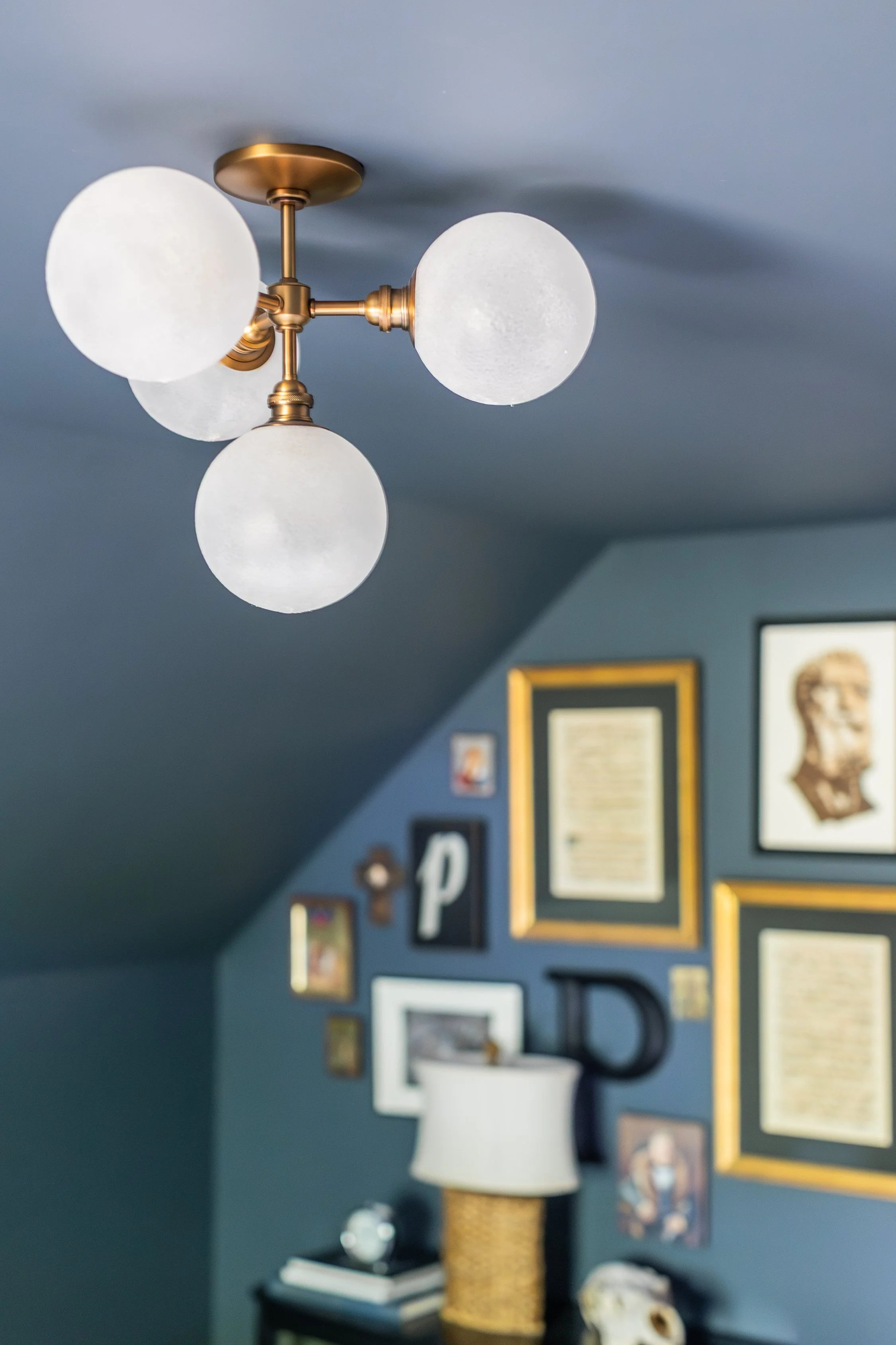 A ceiling light fixture with four spherical frosted glass bulbs attached to a brass ceiling mount in a room with dark blue walls decorated with picture frames and artwork.