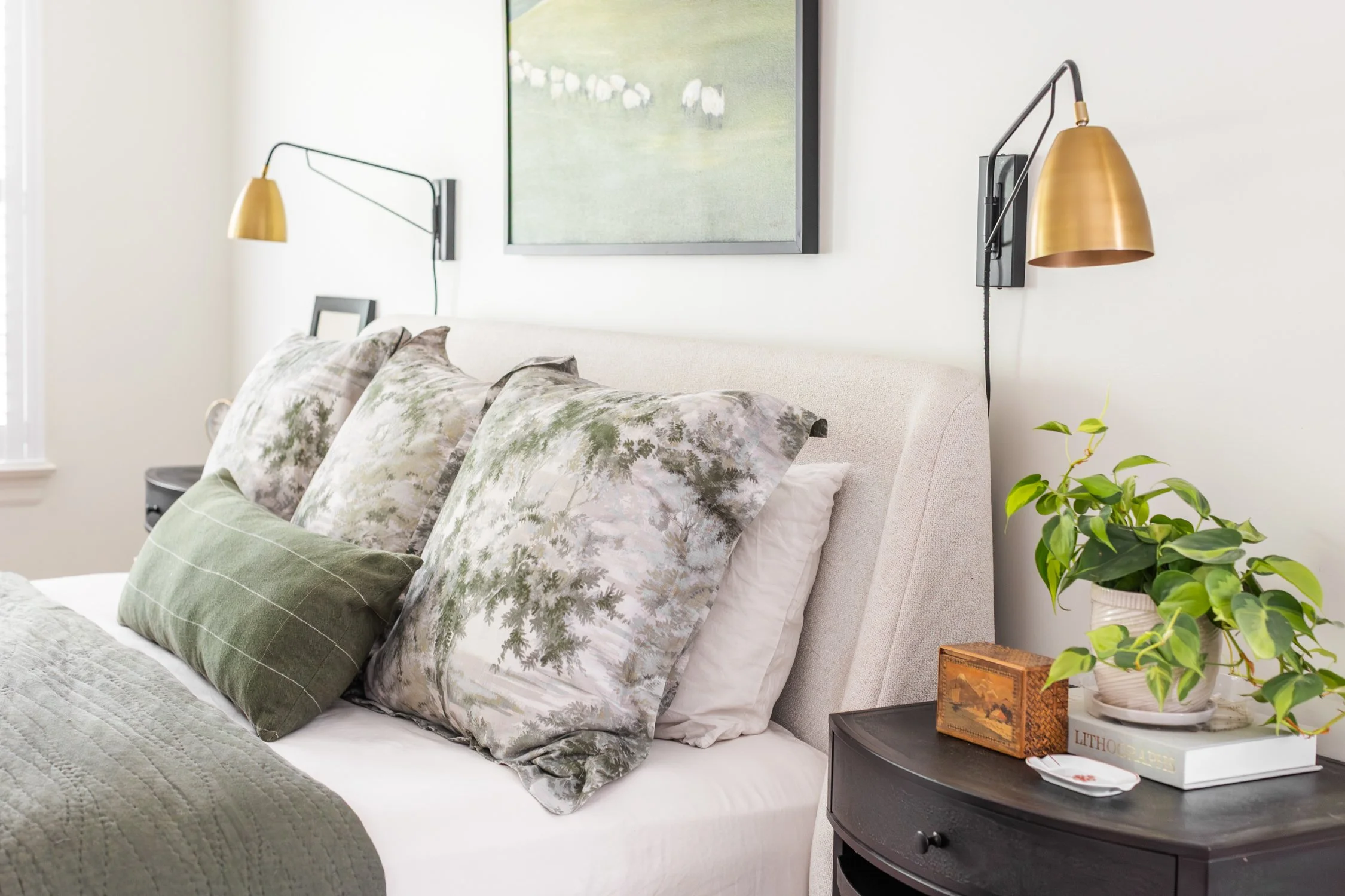 A neatly made bed with pillows featuring nature-themed pillowcases, a bedside table with decorative items, and wall-mounted bedside lamps in a bright, cozy bedroom.