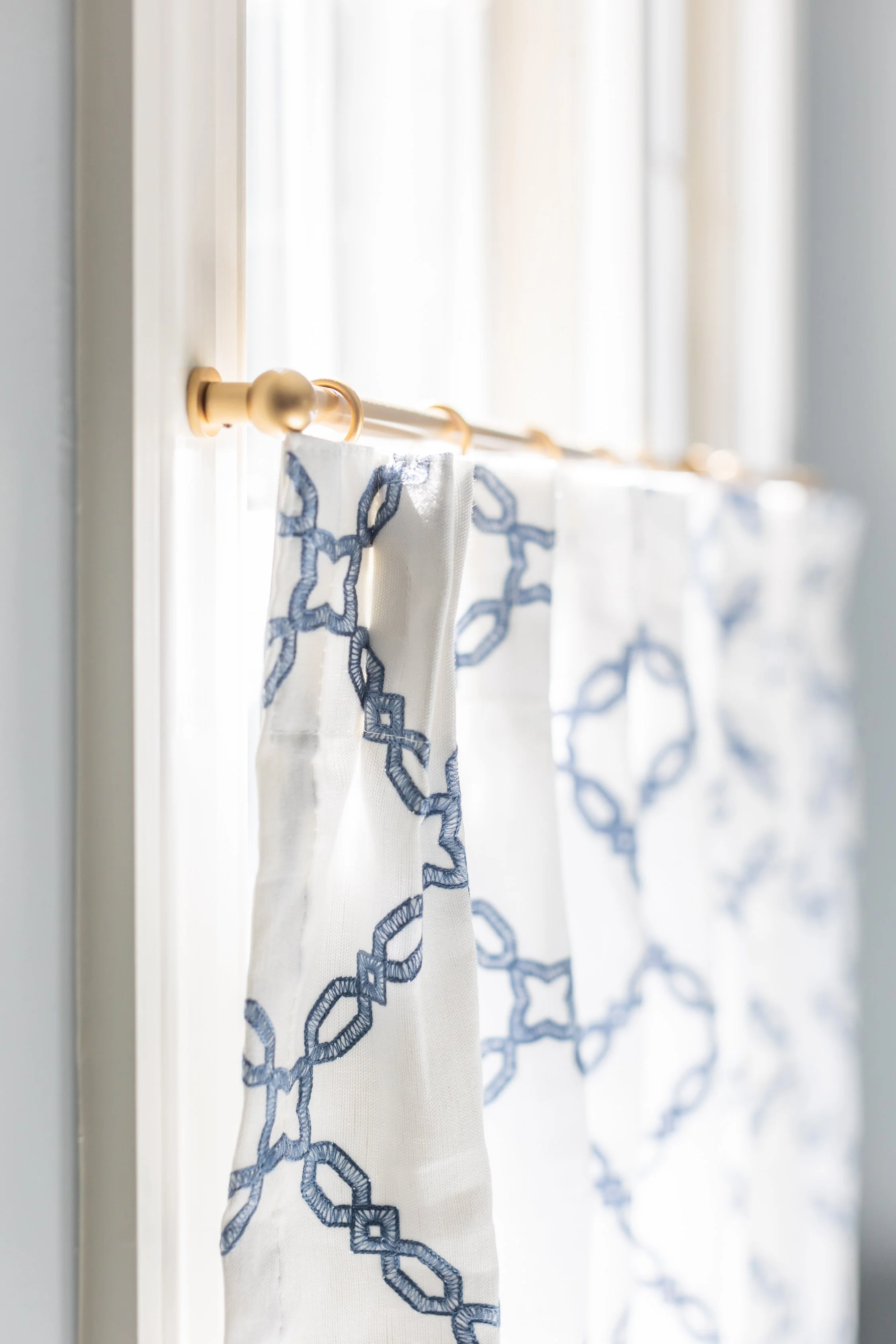 White curtains with blue embroidered chain-link pattern hanging on a gold curtain rod in front of a window.