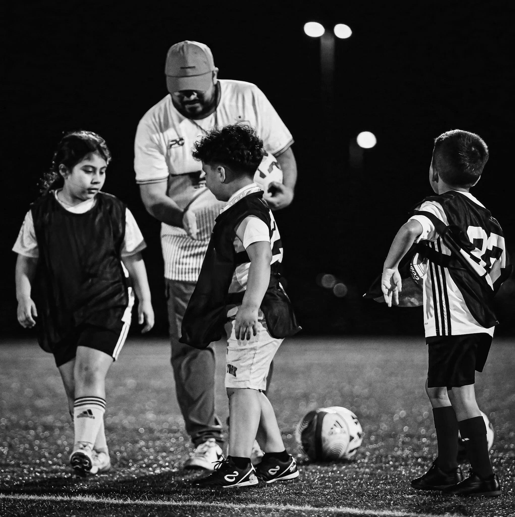 Black and white photograph of young soccer players on a field at night, with a coach or referee supervising.