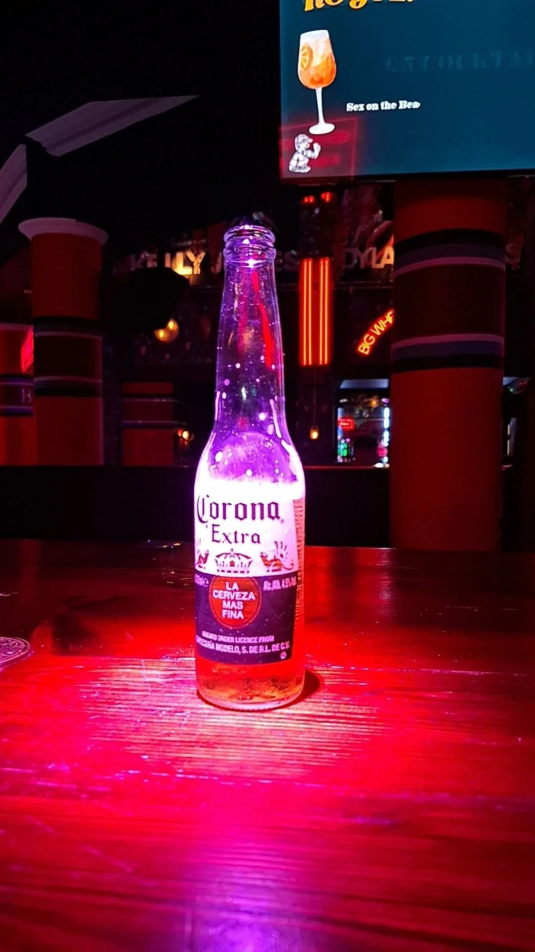 A cold bottle of Corona Extra beer on a wooden table in a bar with red and purple neon lighting.