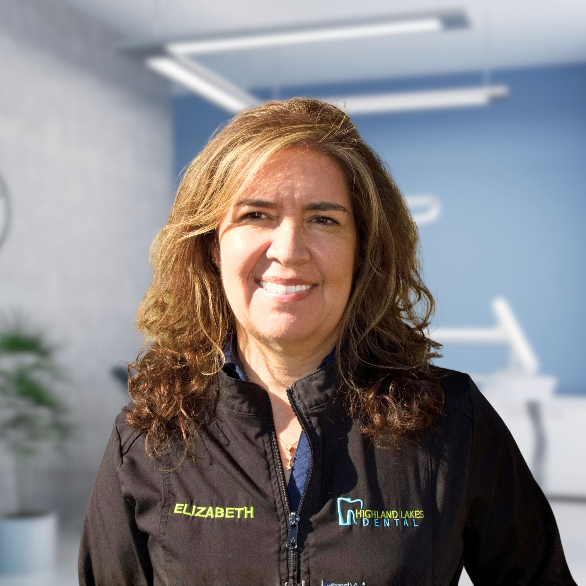 Close-up of a smiling woman with brown hair, wearing a black jacket with the name Elizabeth embroidered in green and a dental office logo for Highland Lakes Dental.