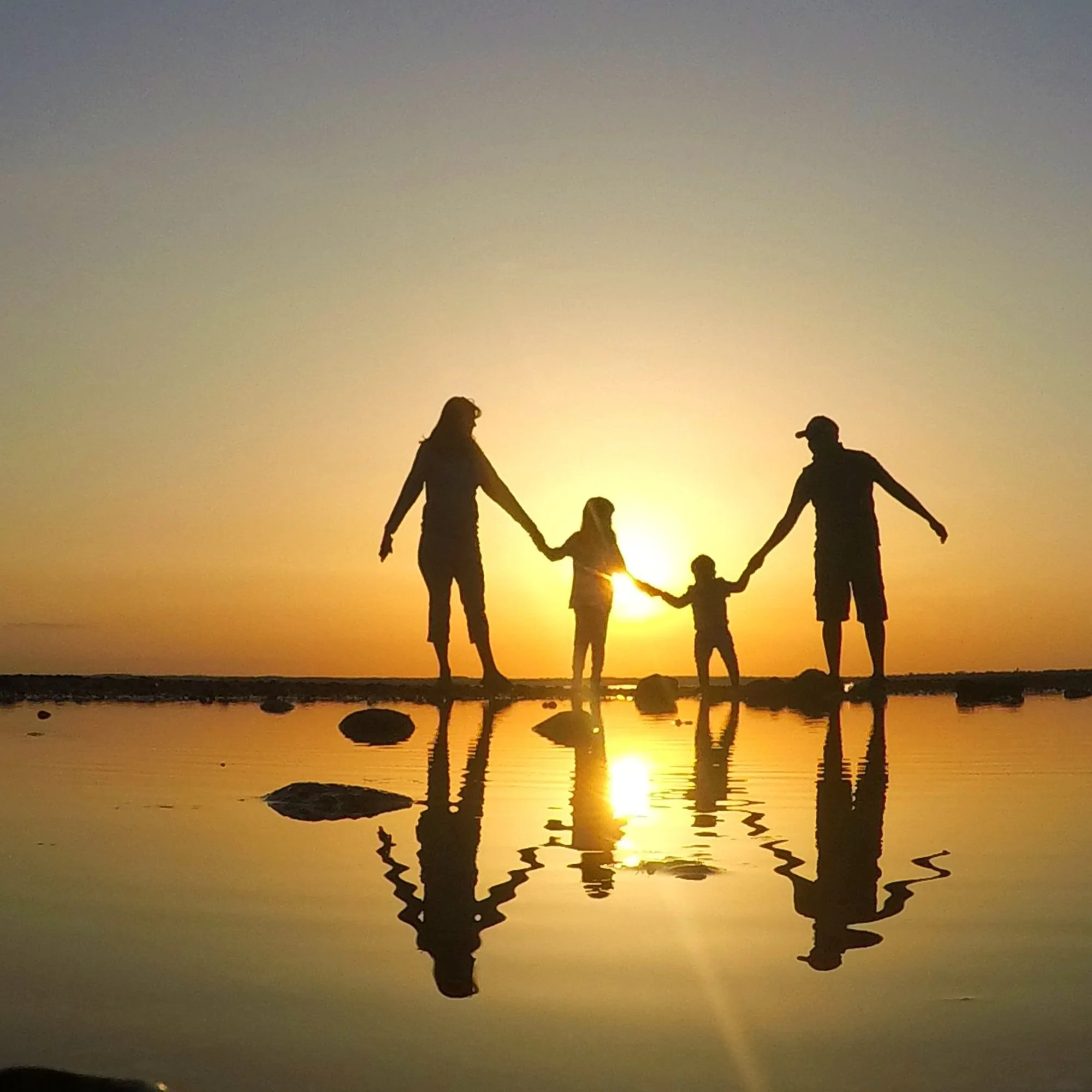 Silhouettes of a family holding hands at sunset near water with rocks, creating reflections.