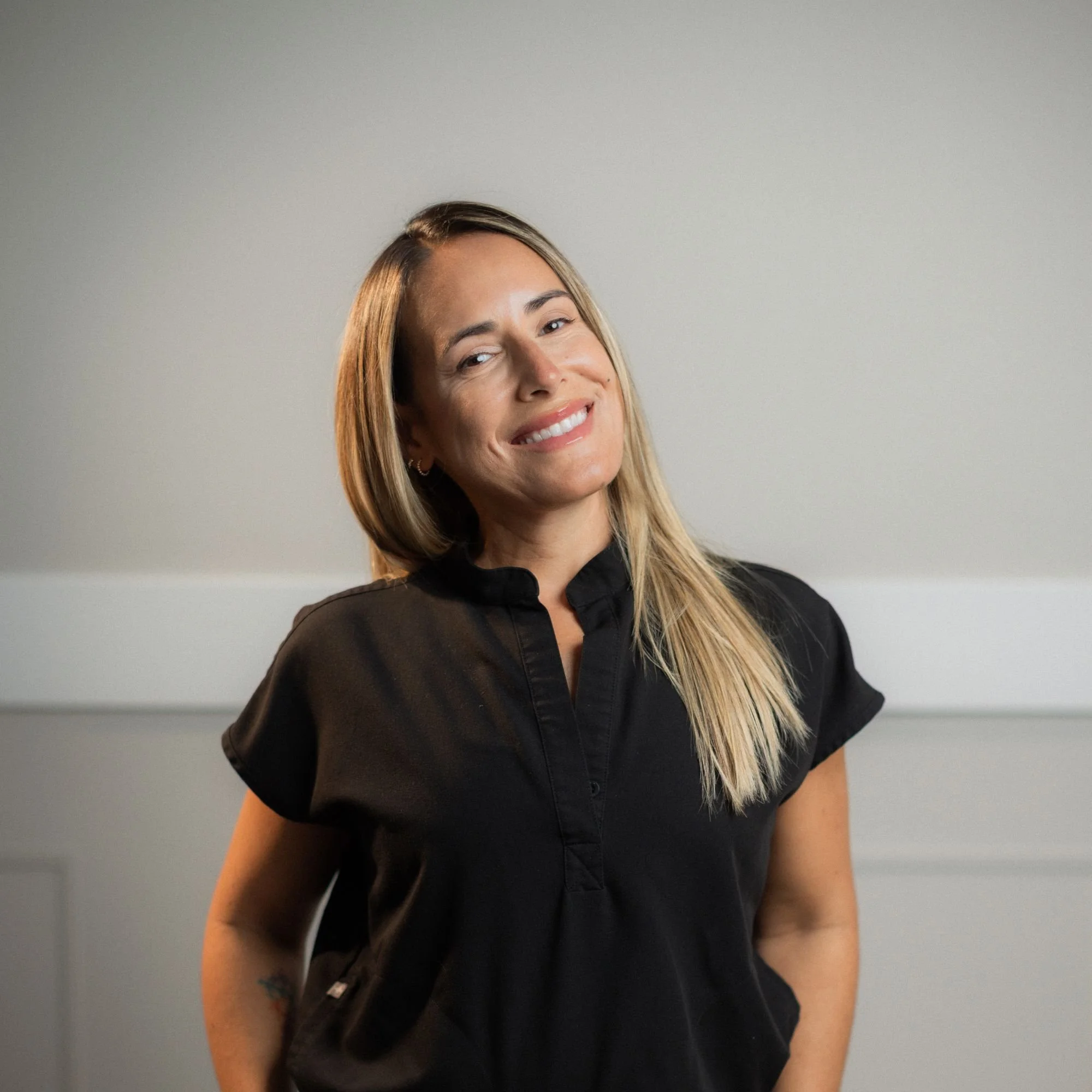 A woman with long, blonde hair wearing a black top grinning at the camera, standing against a plain, light-colored background.