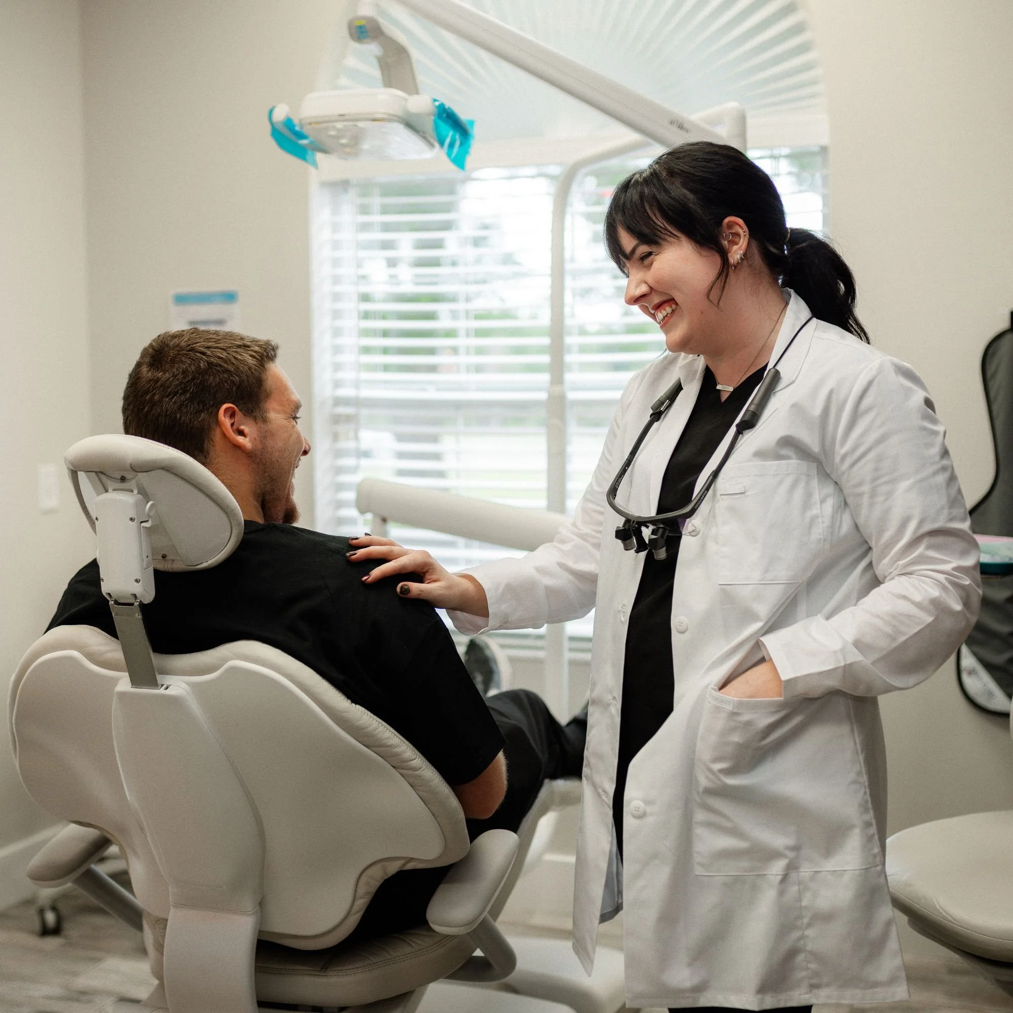 A dentist wearing a white coat and with a stethoscope around her neck is smiling and talking to a male patient who is sitting in a dental chair. The dental office has large windows with blinds and dental equipment visible.