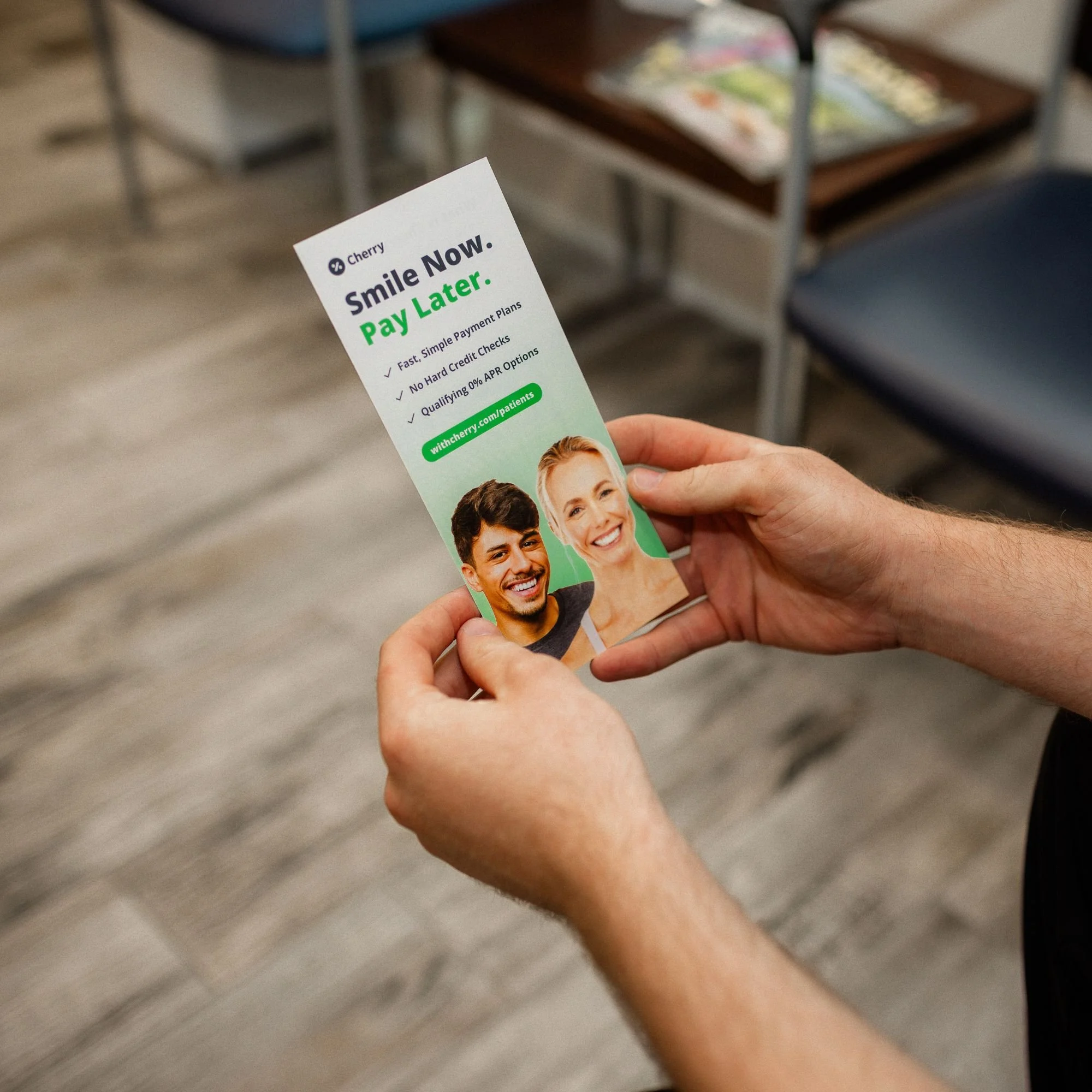 A person holding a promotional flyer for Cherry with the slogan 'Smile Now. Pay Later.' in a room with chairs and magazines.