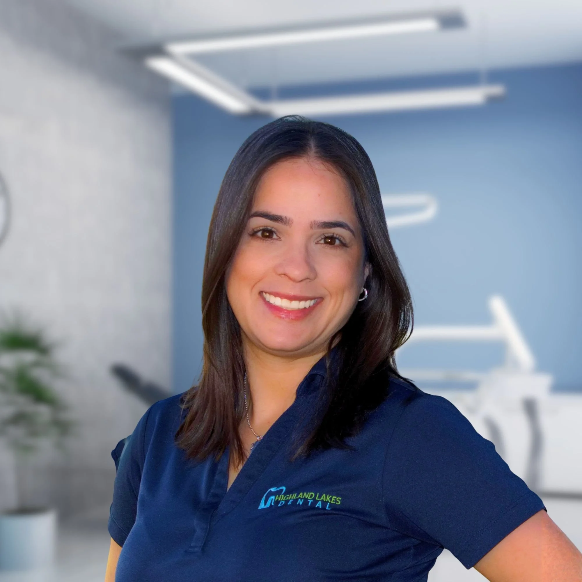 A woman with long dark hair, smiling, wearing a navy blue shirt with the logo 'Highland Lakes Dental' in green and blue. She appears to be in a dental office, with blurred background features including a ceiling light.