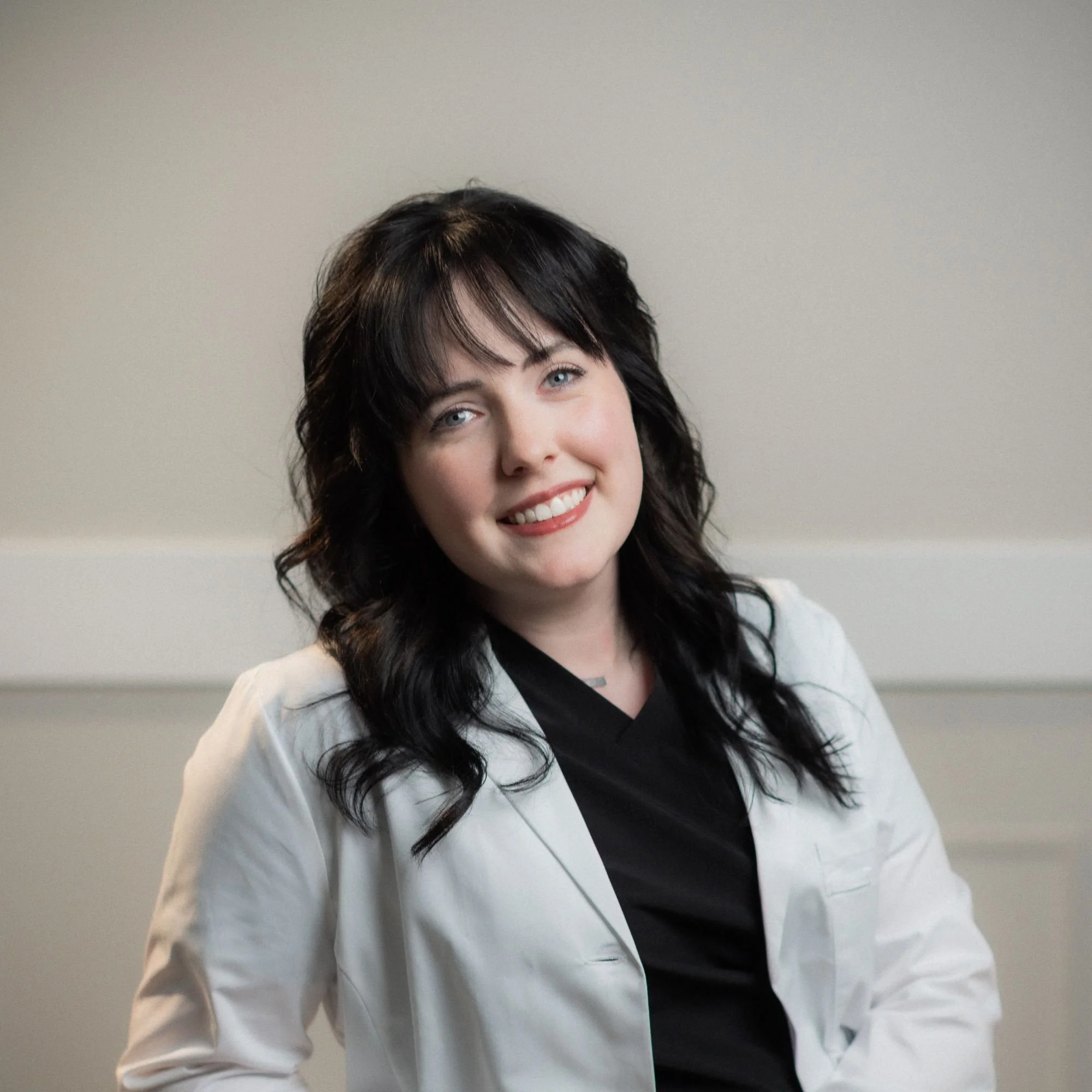Close-up photograph of a woman with dark wavy hair, wearing a white jacket with black inner shirt, smiling, against a plain beige background.