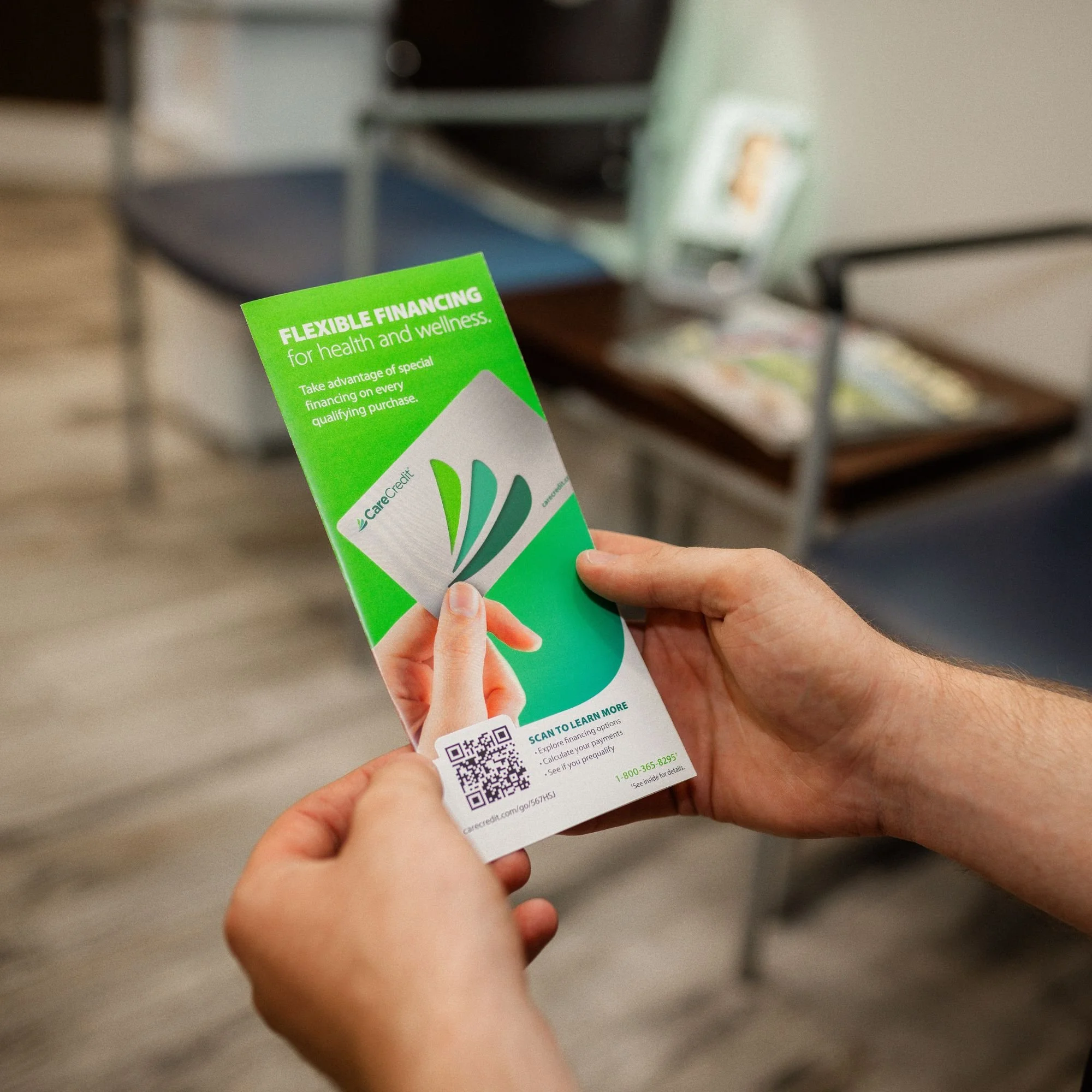 Person holding a green Credit Karma flyer with a QR code, in a room with shelves and furniture in the background