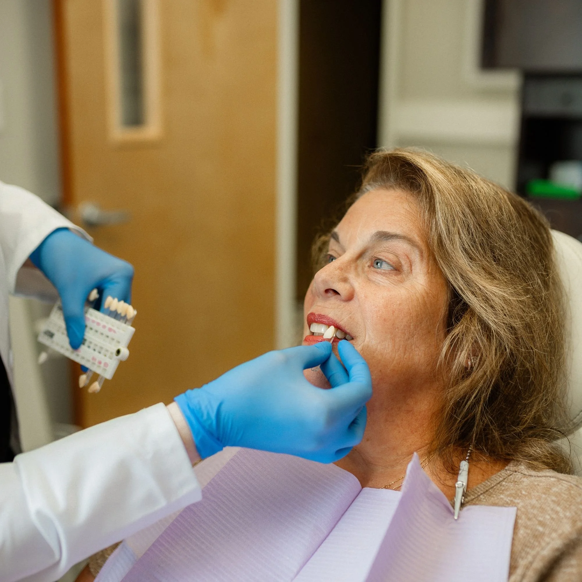 A woman lying in a medical chair, receiving a dental exam or treatment from a healthcare professional wearing blue gloves and a white coat, in a clinic.
