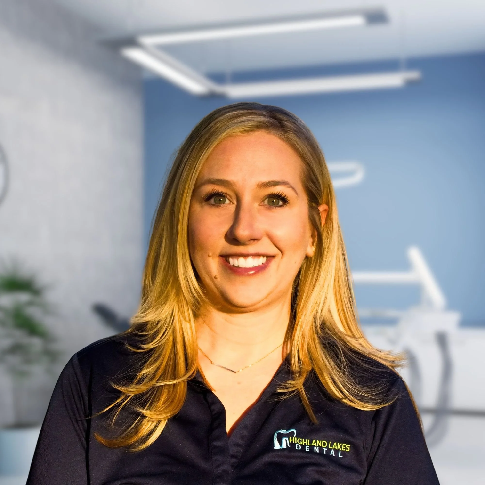 Close-up of a woman smiling, wearing a black shirt with a logo that reads 'Highland Lakes Dental,' with long blonde hair and blue eyes.